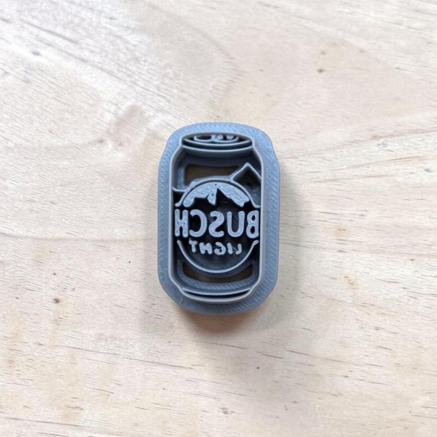 A plastic gray cookie cutter with a logo that reads 'BUSCH LIGHT' and a mountain graphic, placed on a light wood surface.