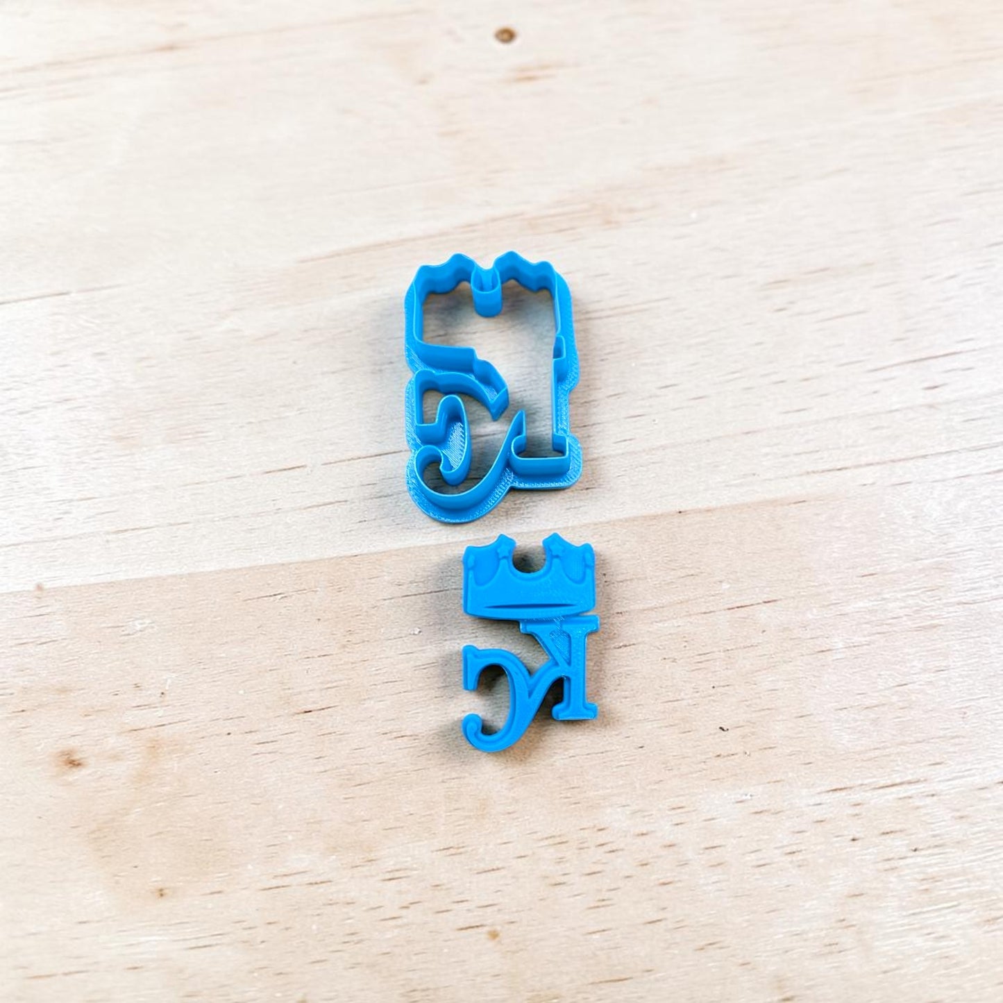 Two blue cookie cutters shaped like stylized letters on a light wooden surface.