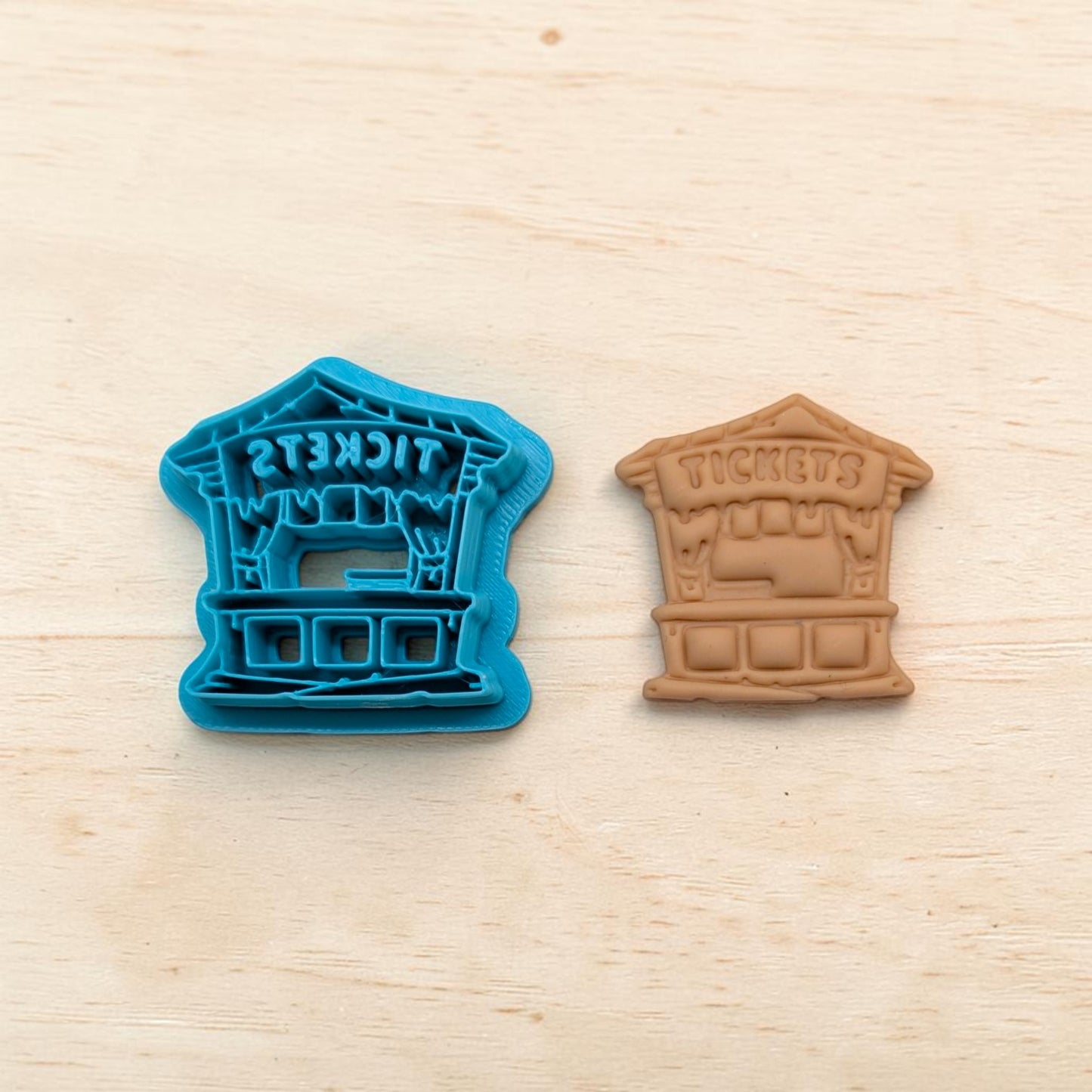 Blue haunted ticket booth cookie cutter beside tan clay imprint showing the word ‘TICKETS’ and spooky booth details on a light wood background.