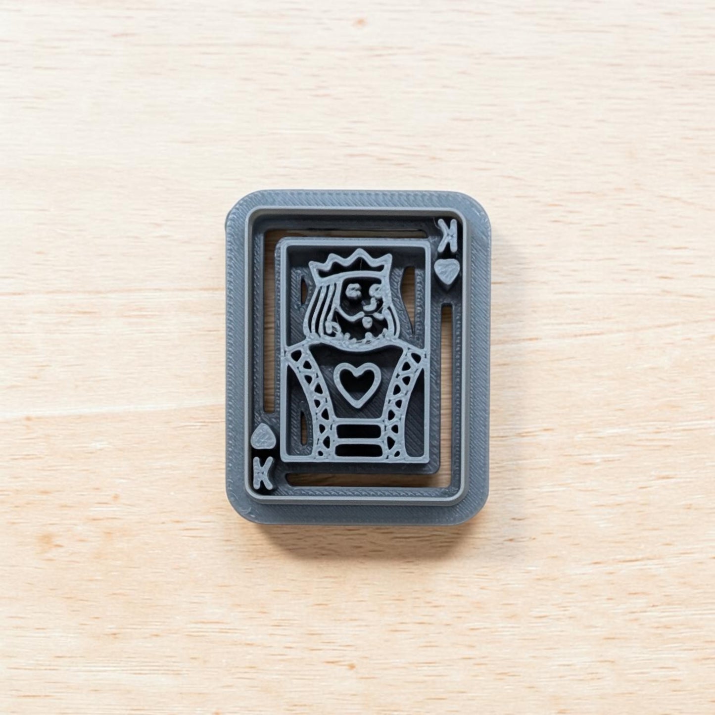 A 3D-printed gray playing card 'King of Hearts' stamp or mold, shown from a top-down view on a light wood grain surface.
