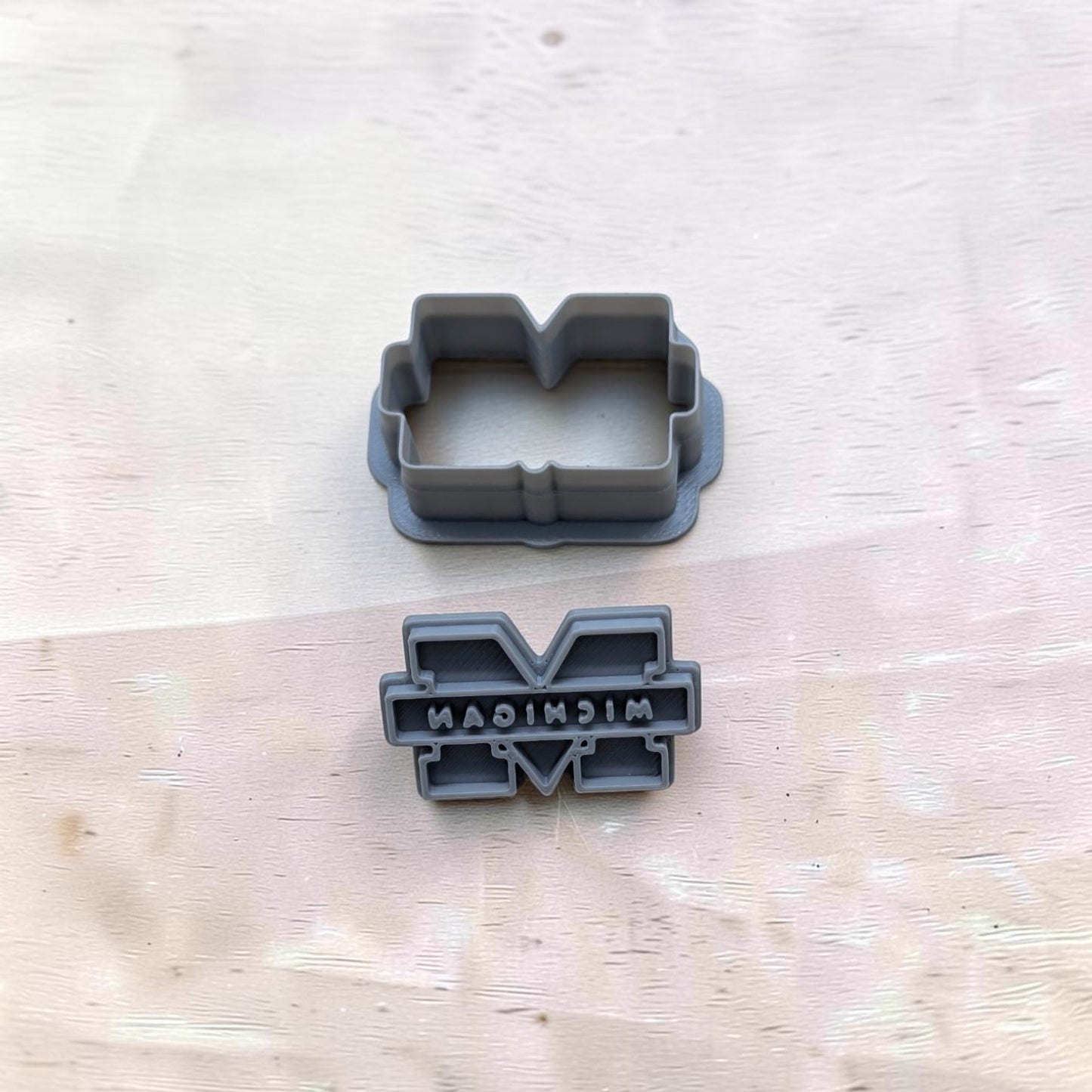 University of Michigan M cookie cutter set with matching embossing stamp — gray 3D printed cutters for cookies, fondant, and crafts on light wood surface.