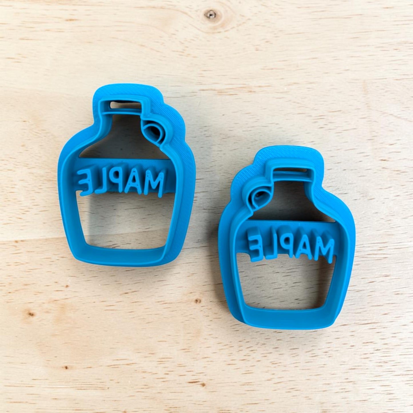 Two bright blue bottle-shaped cookie cutters with the word 'MAPLE' embossed on the inside, displayed on a light wood grain surface.
