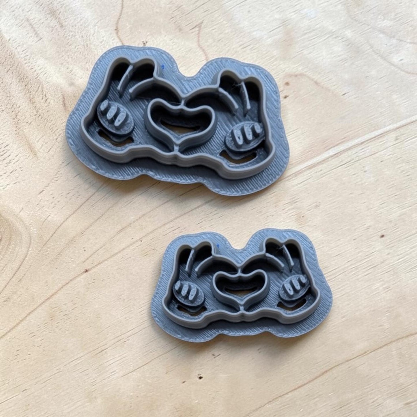 Two gray 3D-printed cookie cutters featuring Mickey Mouse gloved hands forming a heart, shown on a light wood background.