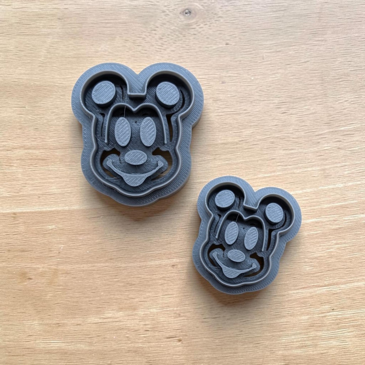 Two dark gray 3D-printed Mickey Mouse-shaped molds on a light wood grain surface.
