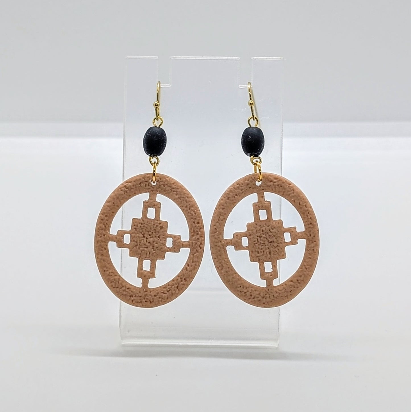 Handmade Dame Dame African symbol earrings with tan geometric cutout pendants and gold hooks displayed on clear stand.