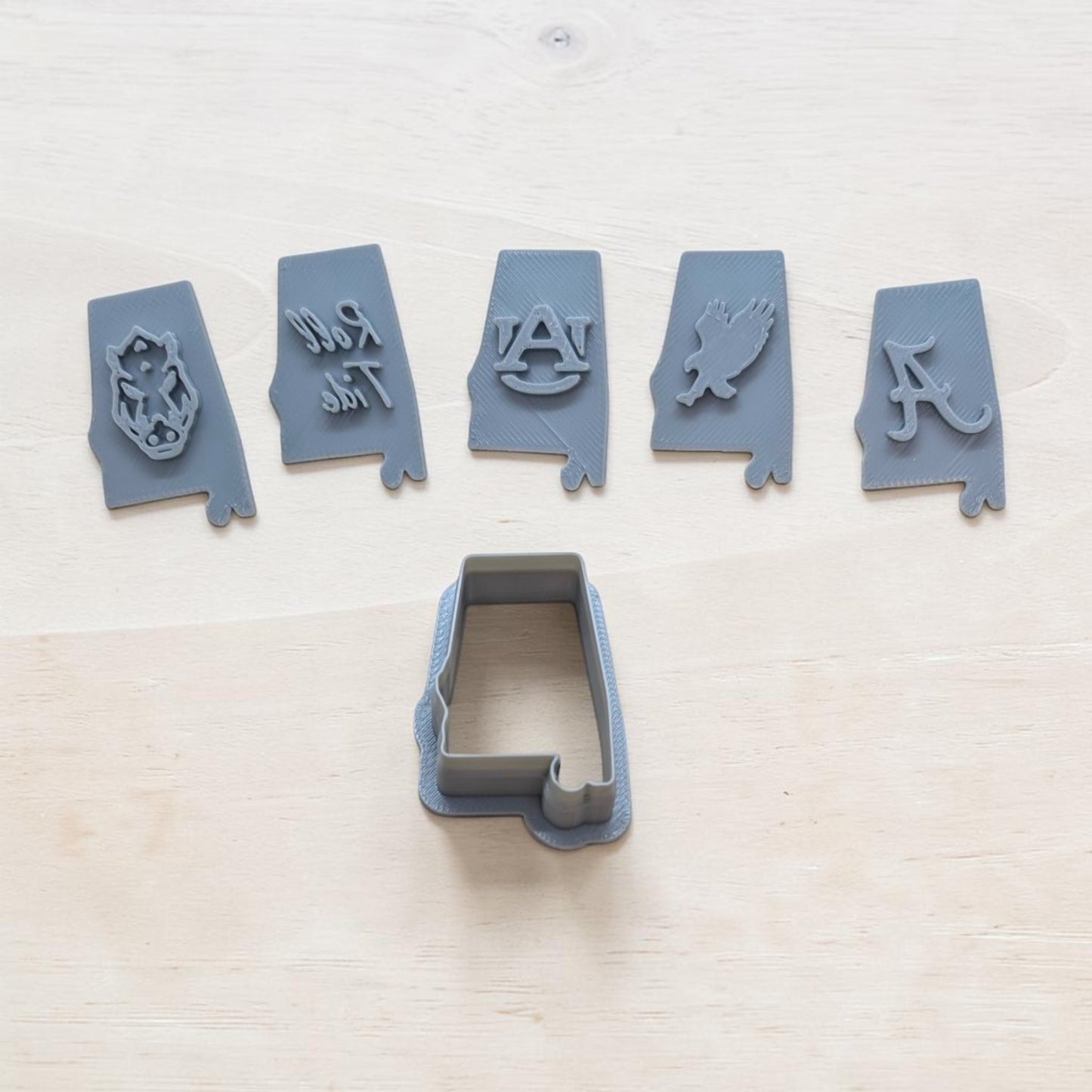 A set of gray cookie cutters arranged on a light wood surface, including five Alabama Crimson Tide themed shapes and one larger rectangular cutter below them.