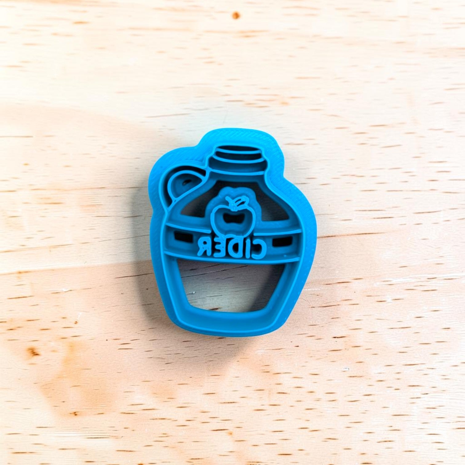 A bright blue plastic apple cider punch cookie cutter with an embossed apple and the word 'CIDER', shown on a light-colored wooden surface.