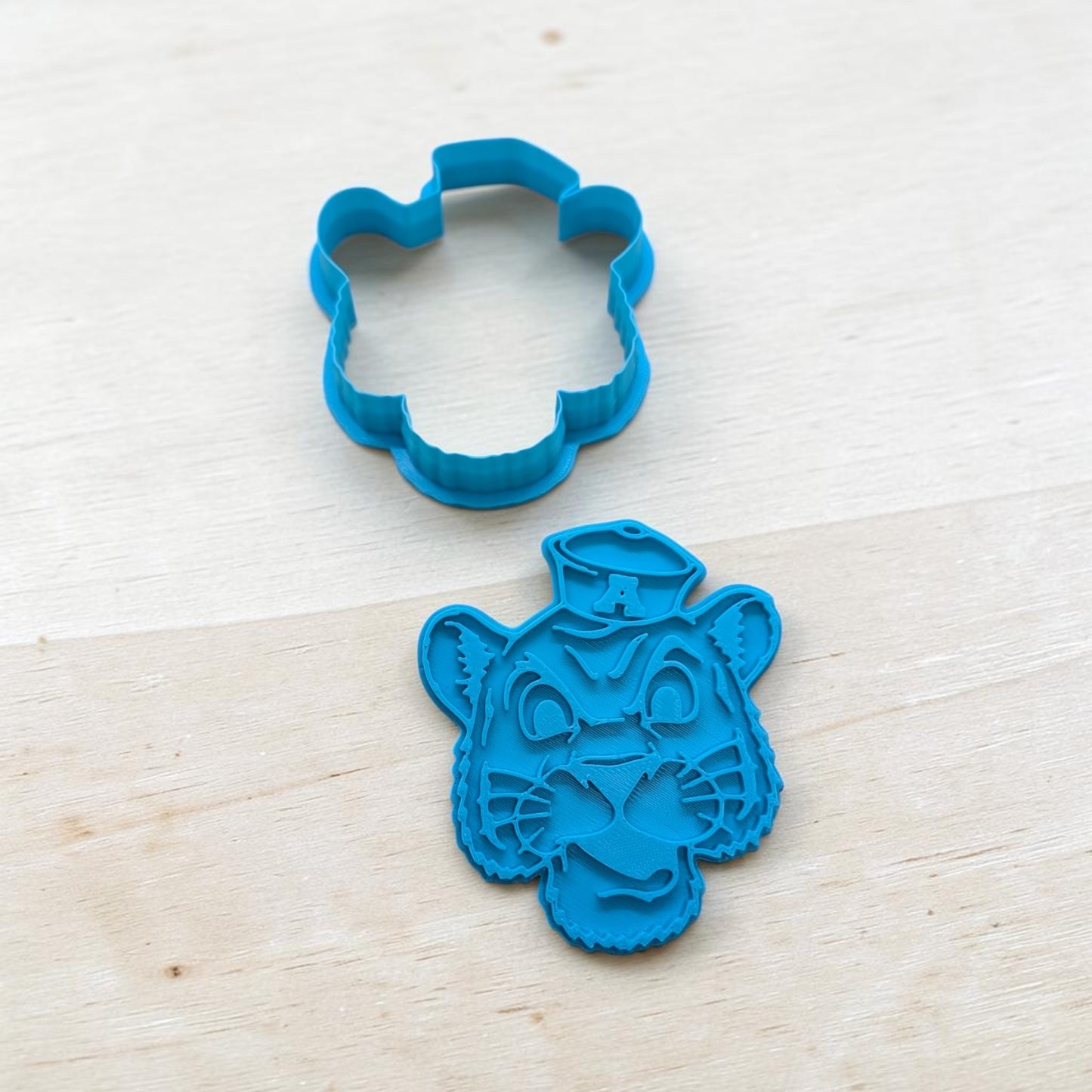 Auburn Tiger cookie cutter and stamp set – blue 3D‑printed outline and detailed embosser for baking, ceramics, and fondant crafts on wood background.