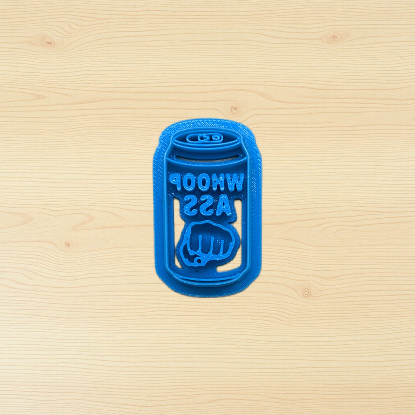 Blue plastic can of whoop ass cookie cutter shaped like a beer can on a wooden surface