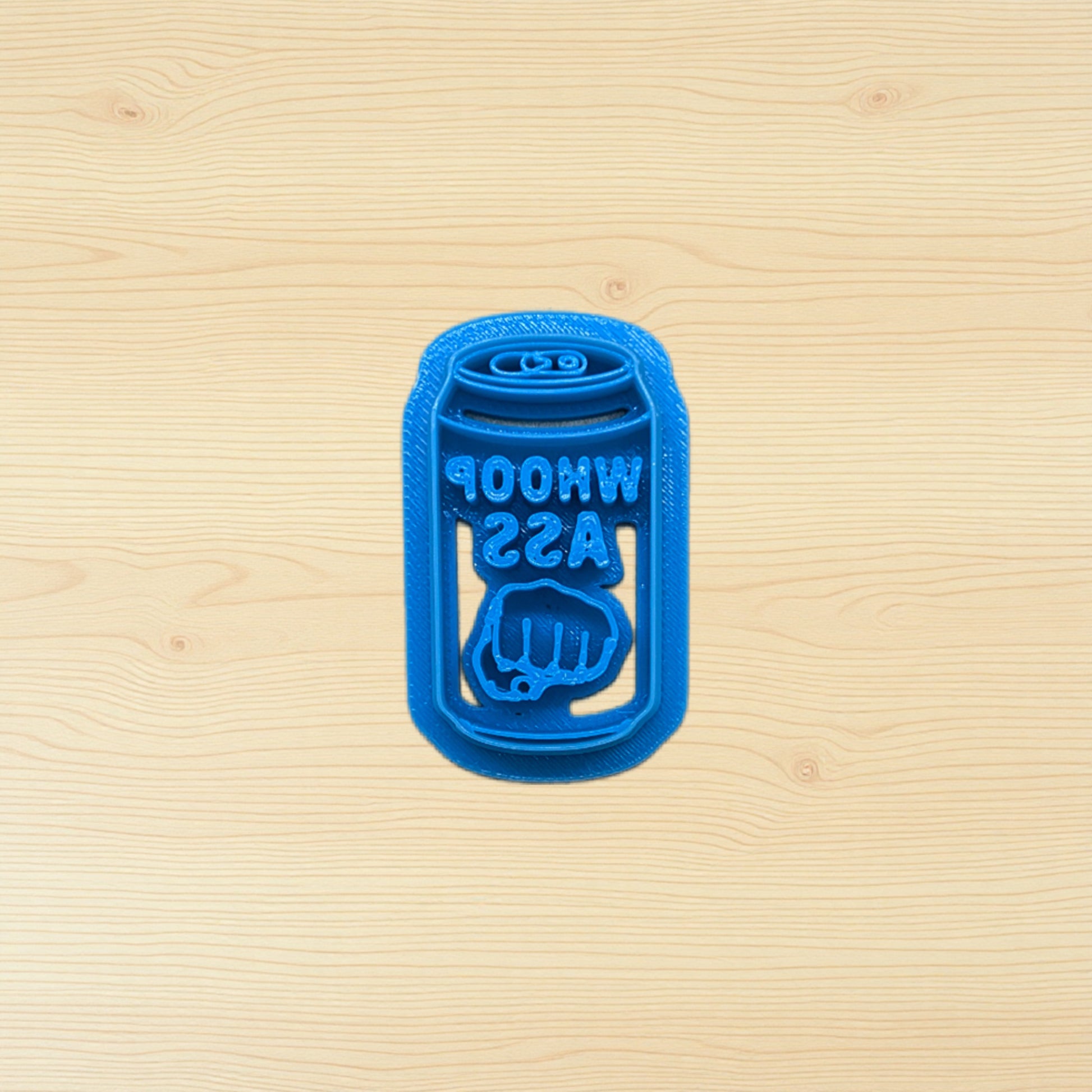 Blue plastic can of whoop ass cookie cutter shaped like a beer can on a wooden surface