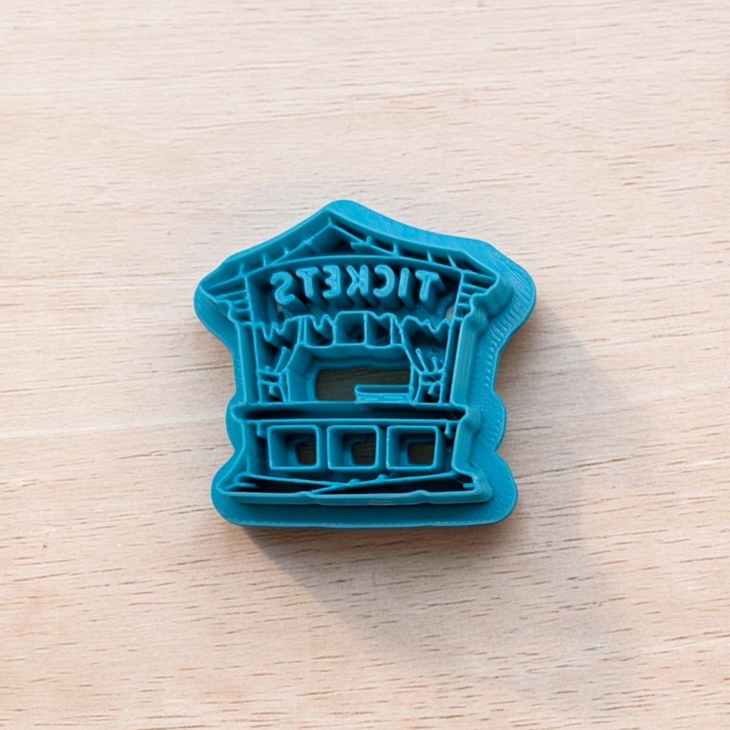 Teal cookie cutter shaped like a haunted carnival ticket booth with icicle‑style drips and reversed ‘TICKETS’ text, placed on a light wooden surface.