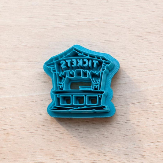 Teal cookie cutter shaped like a haunted carnival ticket booth with icicle‑style drips and reversed ‘TICKETS’ text, placed on a light wooden surface.