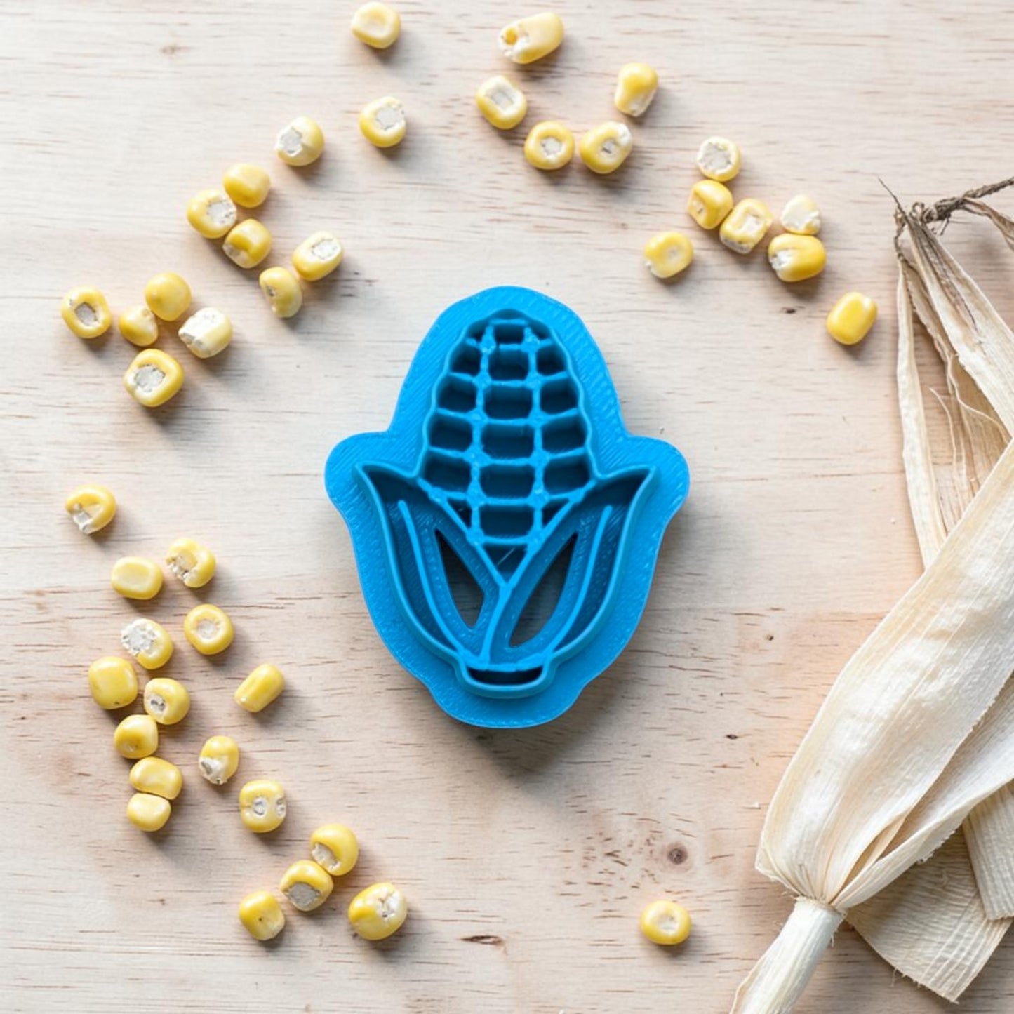 Blue corn-shaped cookie cutter with scattered yellow corn kernels on a wooden surface.