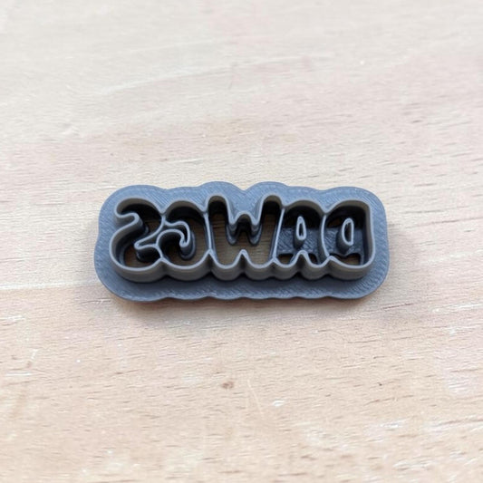A dark gray 3D-printed cookie cutter with the word 'DAWGS' cut into the bottom half, displayed on a light wood grain surface.
