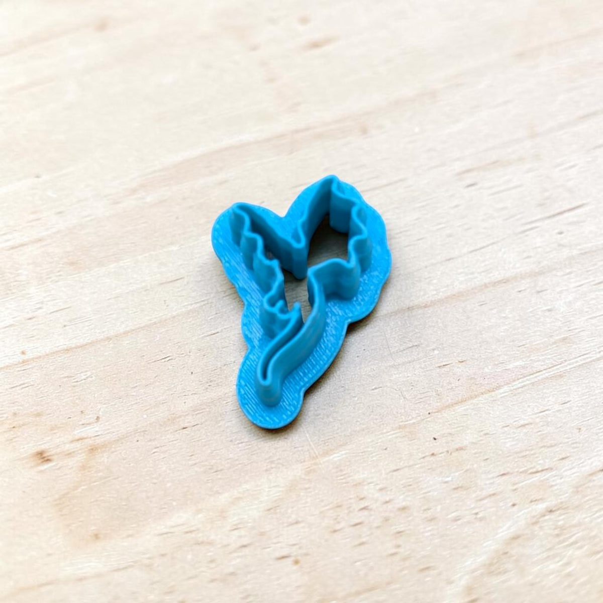 Blue 3D-printed Dragon Silhouette Style C cookie cutter featuring pointed wings and a flowing curved tail, photographed on light wood for fantasy cookies, polymer clay earrings, fondant and crafts.