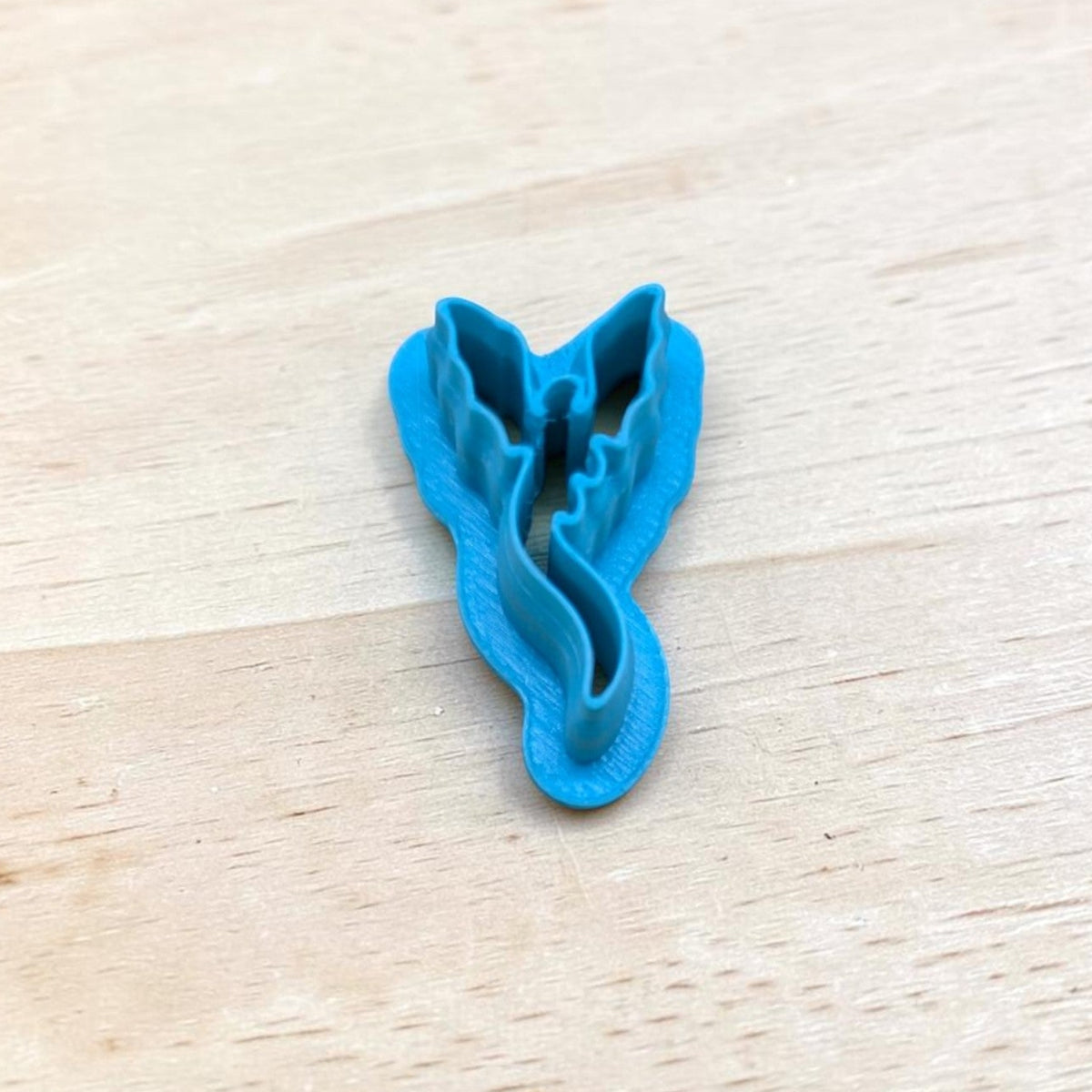 Blue 3D-printed Dragon Silhouette Style B cookie cutter featuring upright wings and a long curved tail, photographed on light wood for fantasy cookies, polymer clay earrings, fondant and crafts.