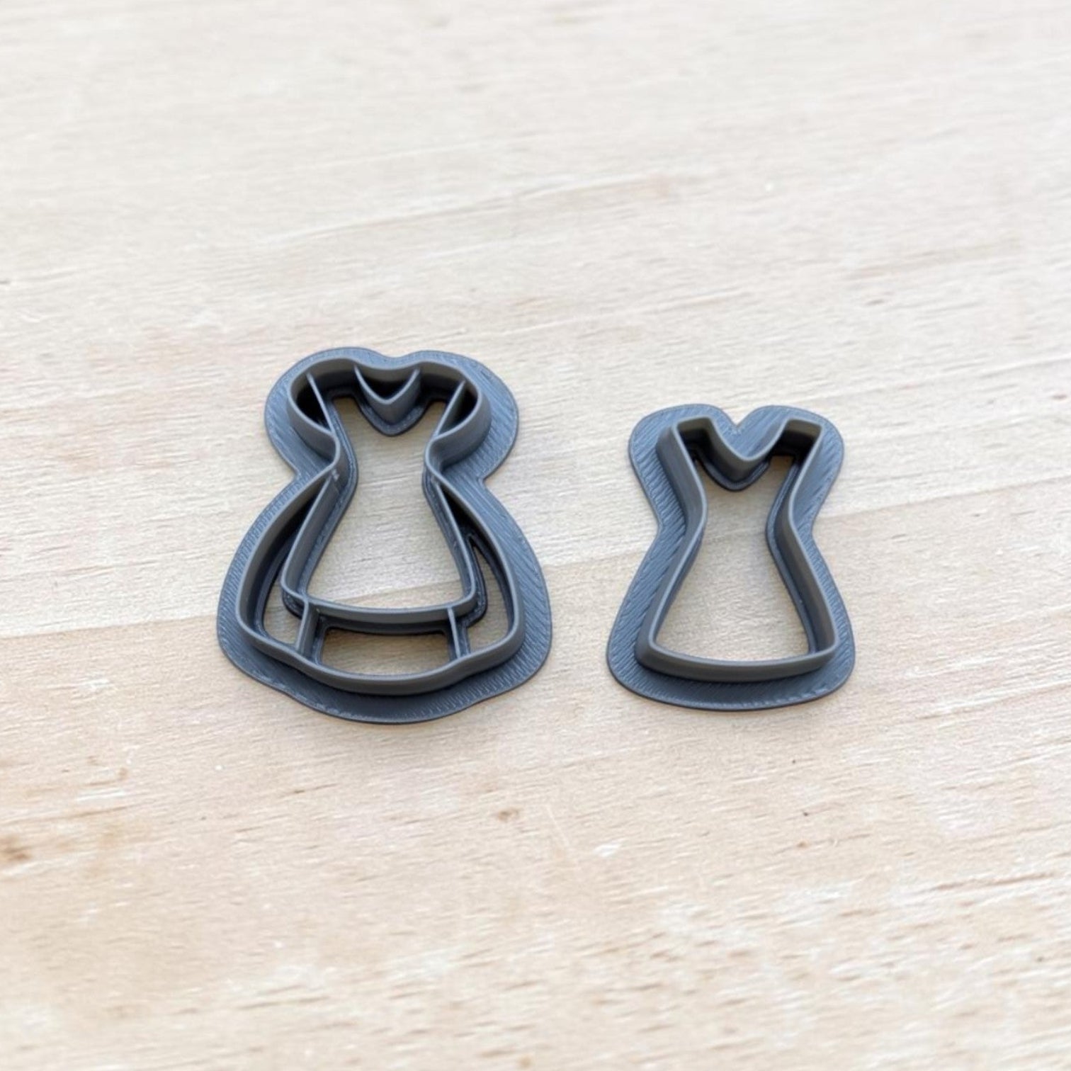 Two dark gray dress-shaped cookie cutters nested together and a smaller standalone cutter, displayed on a light wood grain surface.