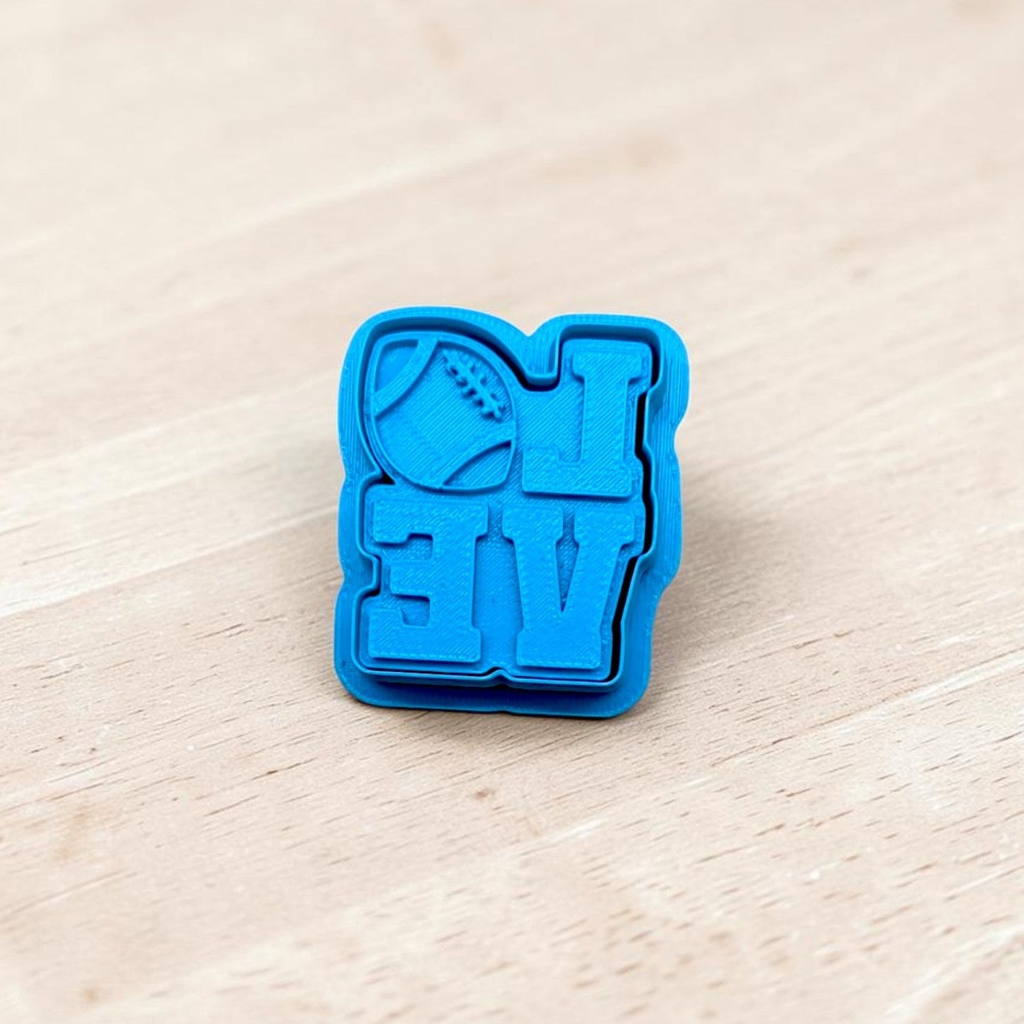 A bright blue plastic cookie cutter with a football and the letters 'LOVE' embossed, resting on a light-colored wooden surface.