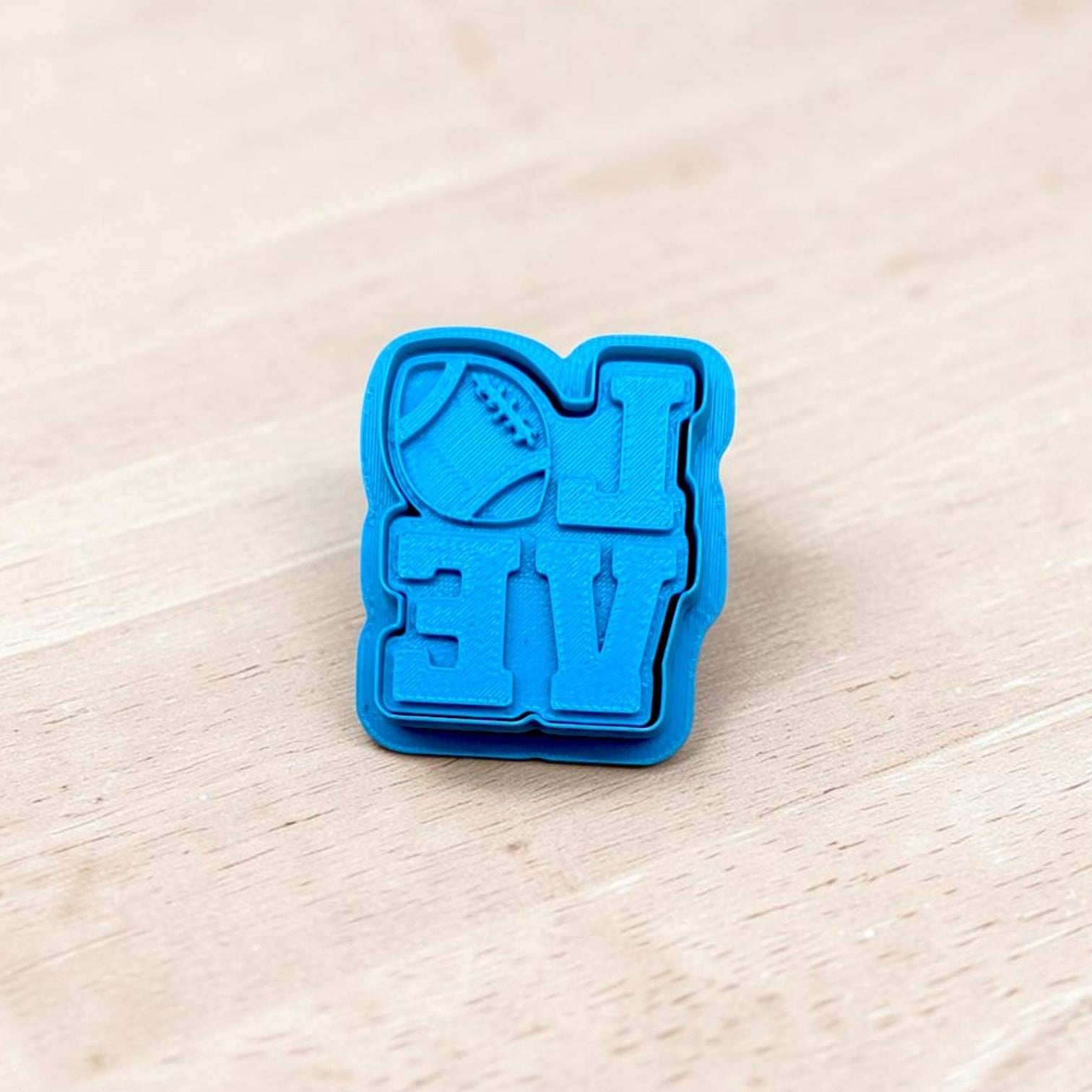 A bright blue plastic cookie cutter with a football and the letters 'LOVE' embossed, resting on a light-colored wooden surface.