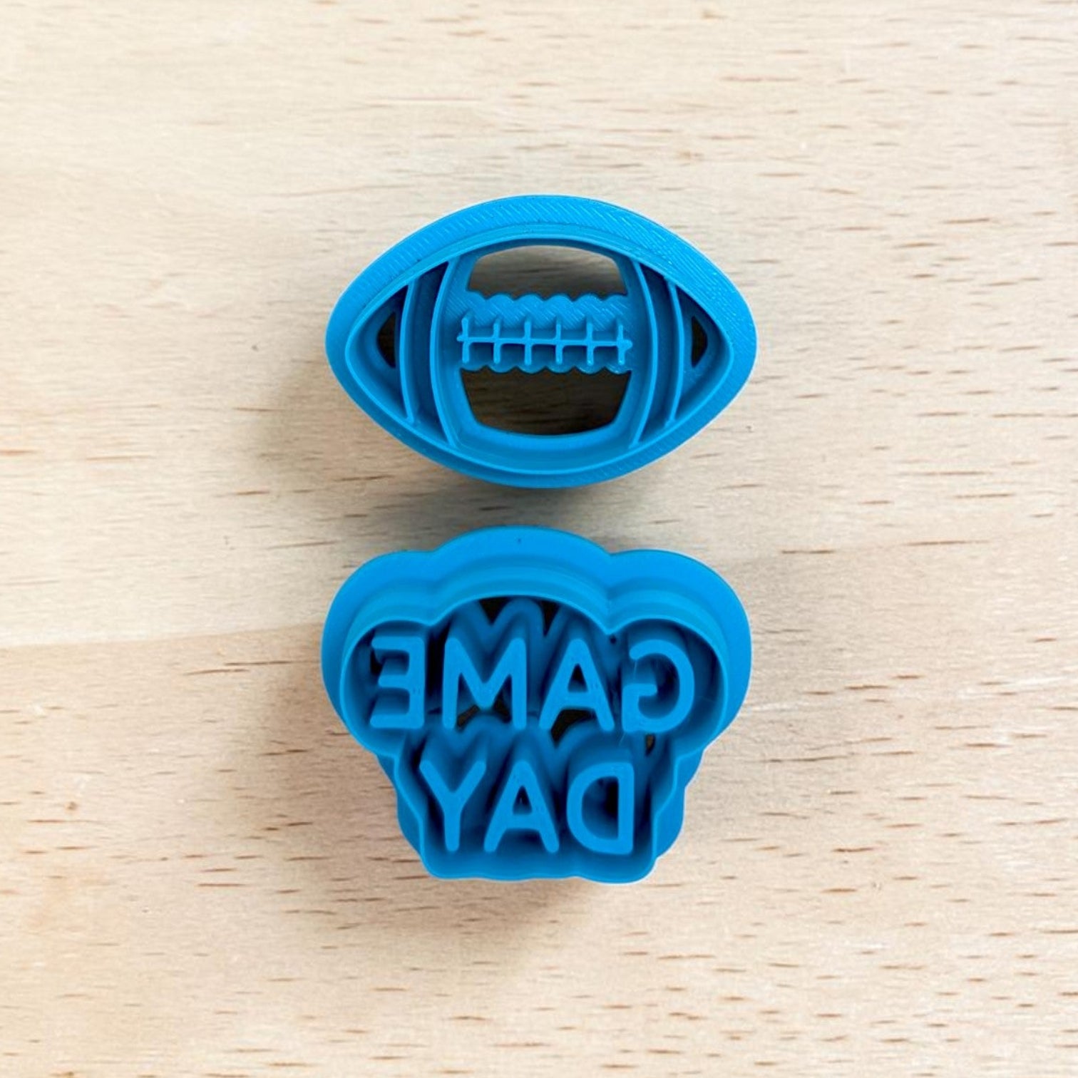 A bright blue plastic cookie cutter set on a light wood background. A football-shaped cutter and a cutter that reads 'GAME DAY'.