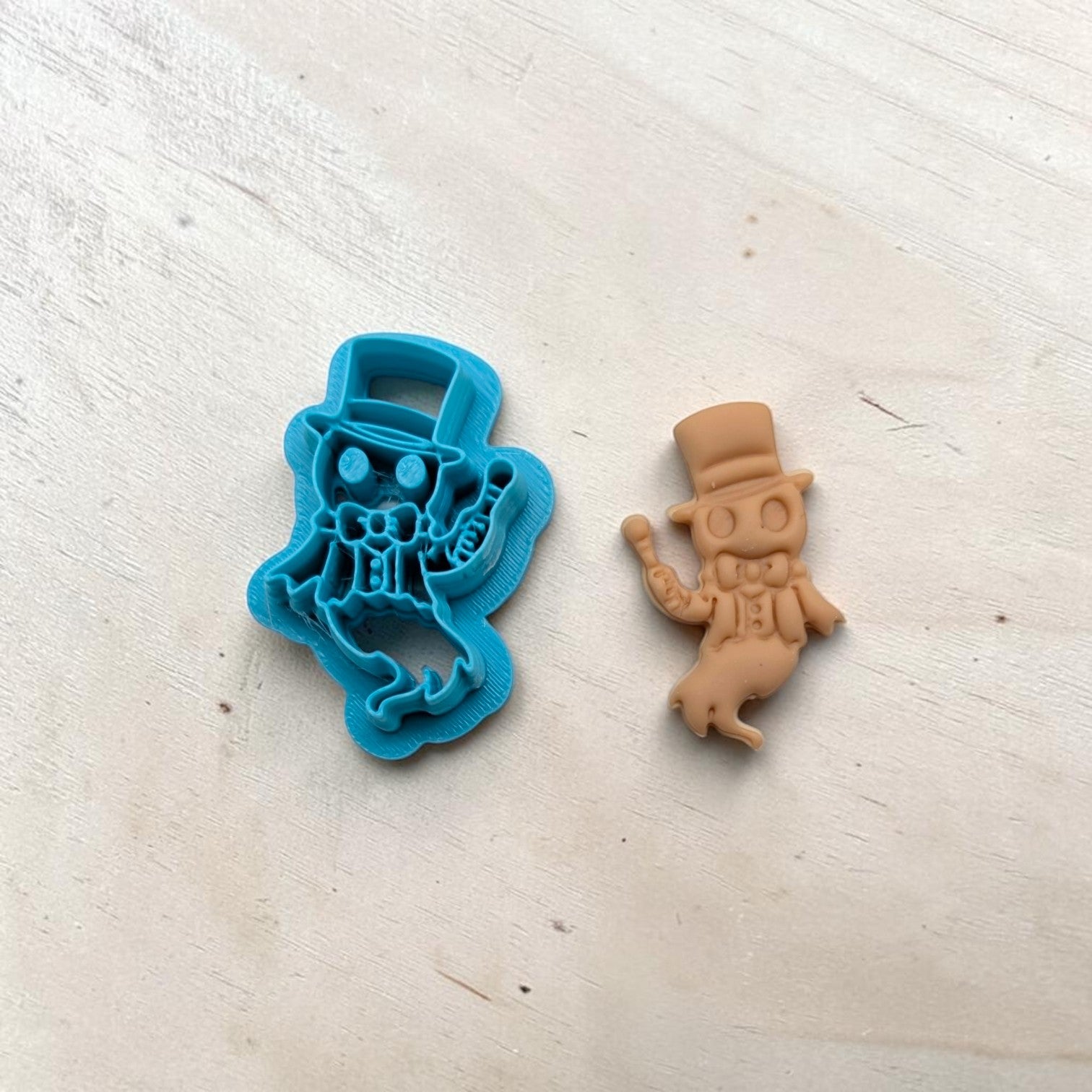 Blue ringmaster ghost cookie cutter beside tan clay imprint showing detailed top hat, bow tie, and cane design.