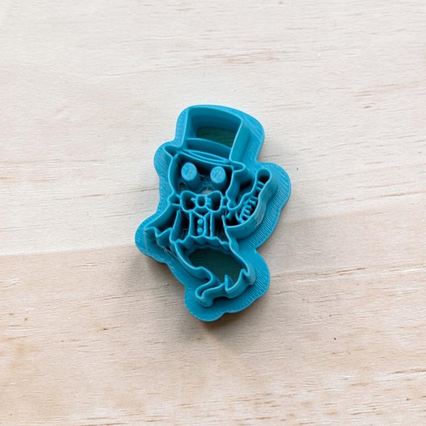 Blue cookie cutter shaped like a ghostly ringmaster wearing a top hat and bow tie, holding a cane, on a light wooden surface.