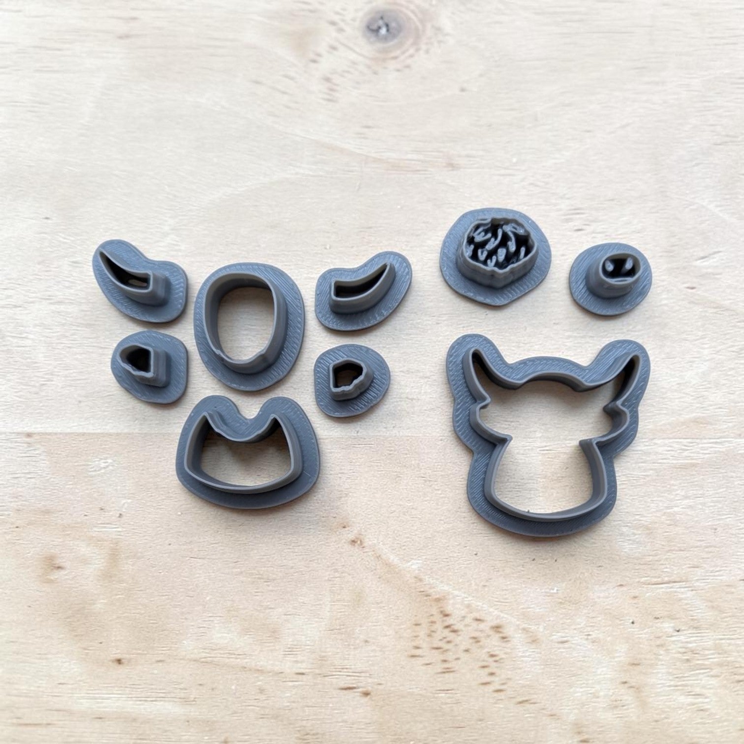A set of dark gray 3D-printed cookie cutters arranged on a light wood grain surface. The set includes various animal and abstract shapes, with the largest piece on the right being a fox head outline cutter.