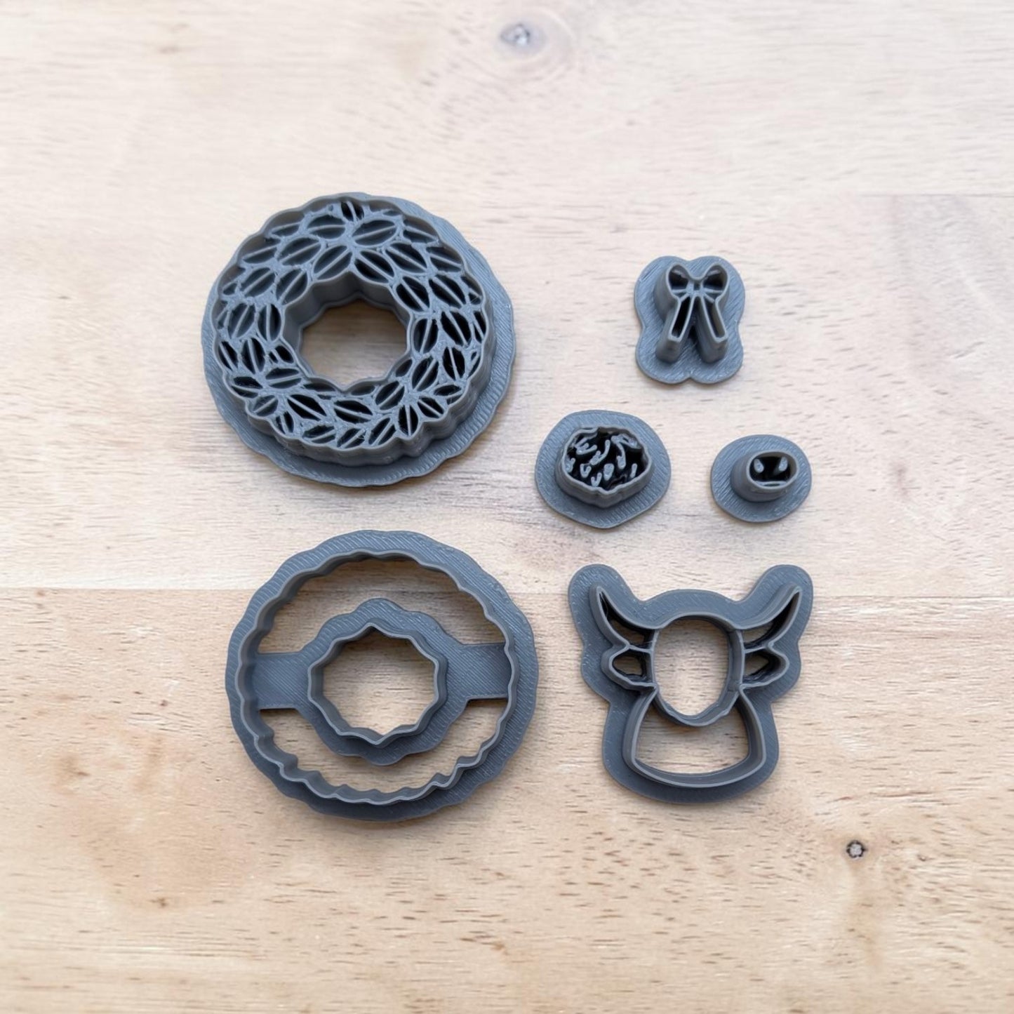 A set of dark gray plastic cookie cutters arranged on a light wood surface, including a floral wreath cutter, a ring cutter, a bow cutter, a floral scroll cutter, and a bull head cutter.