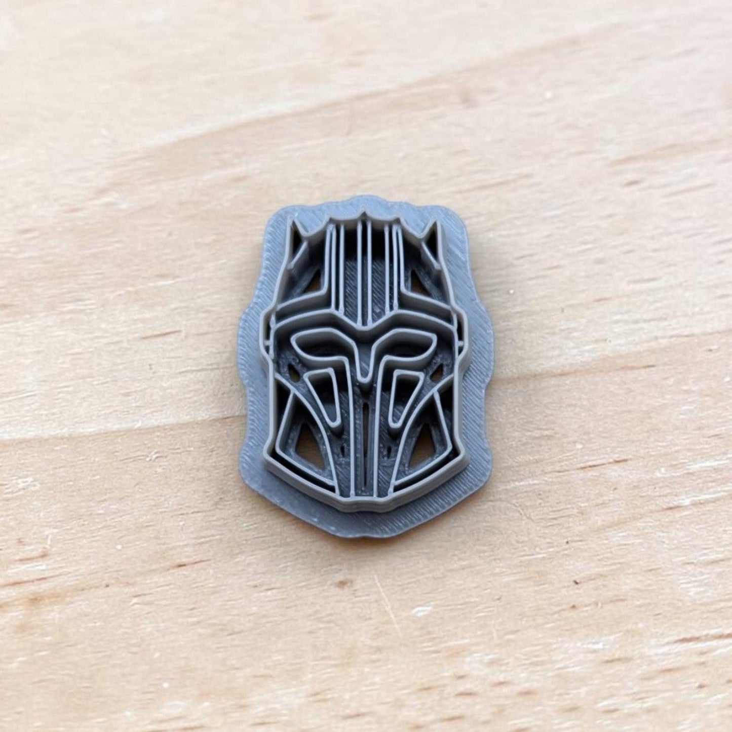 gray 3D printed Star Wars Mandalorian helmet design on wooden surface, detailed geometric pattern, small artisan prototype