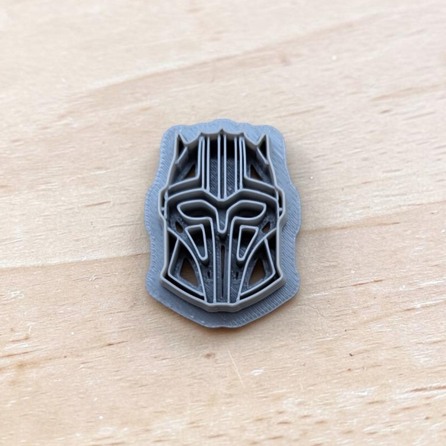 gray 3D printed Star Wars Mandalorian helmet design on wooden surface, detailed geometric pattern, small artisan prototype