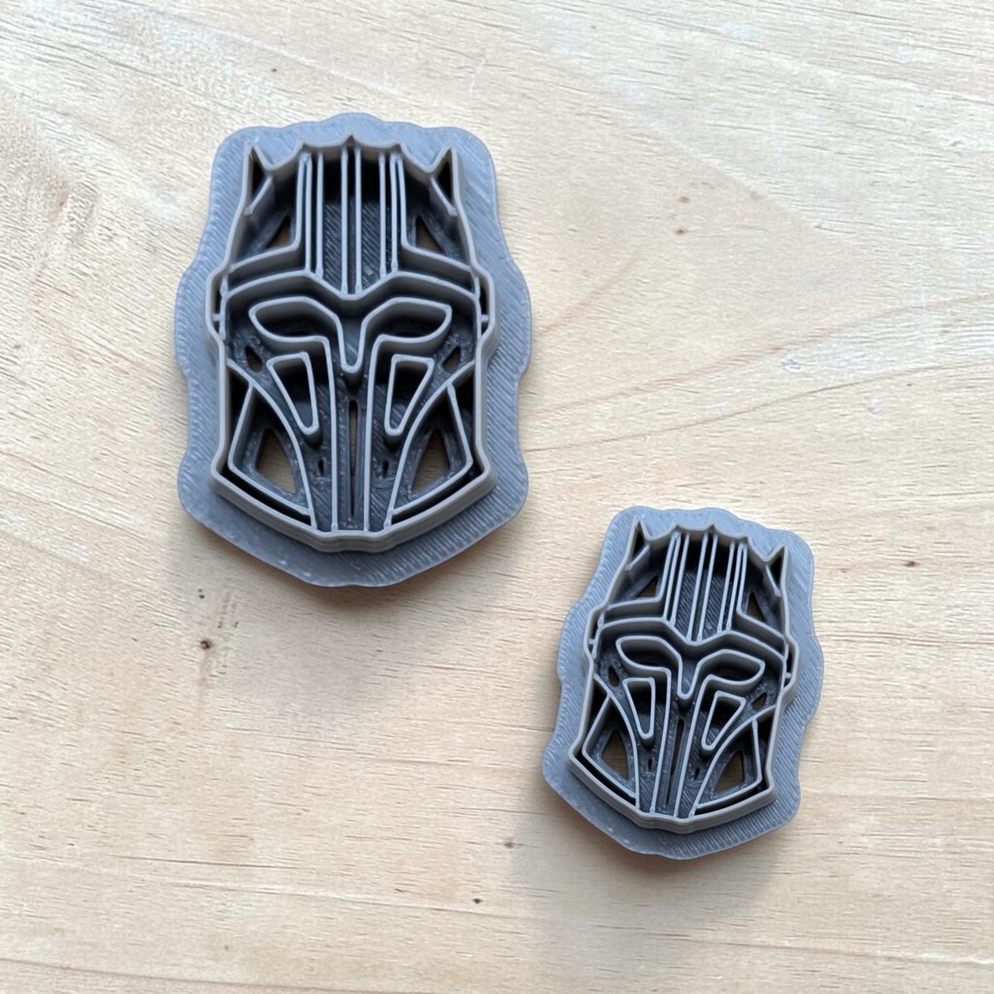 pair of gray 3D printed Star Wars Black Series Mandalorian helmets in different sizes on wood table, angular geometric contours, precision craftsmanship