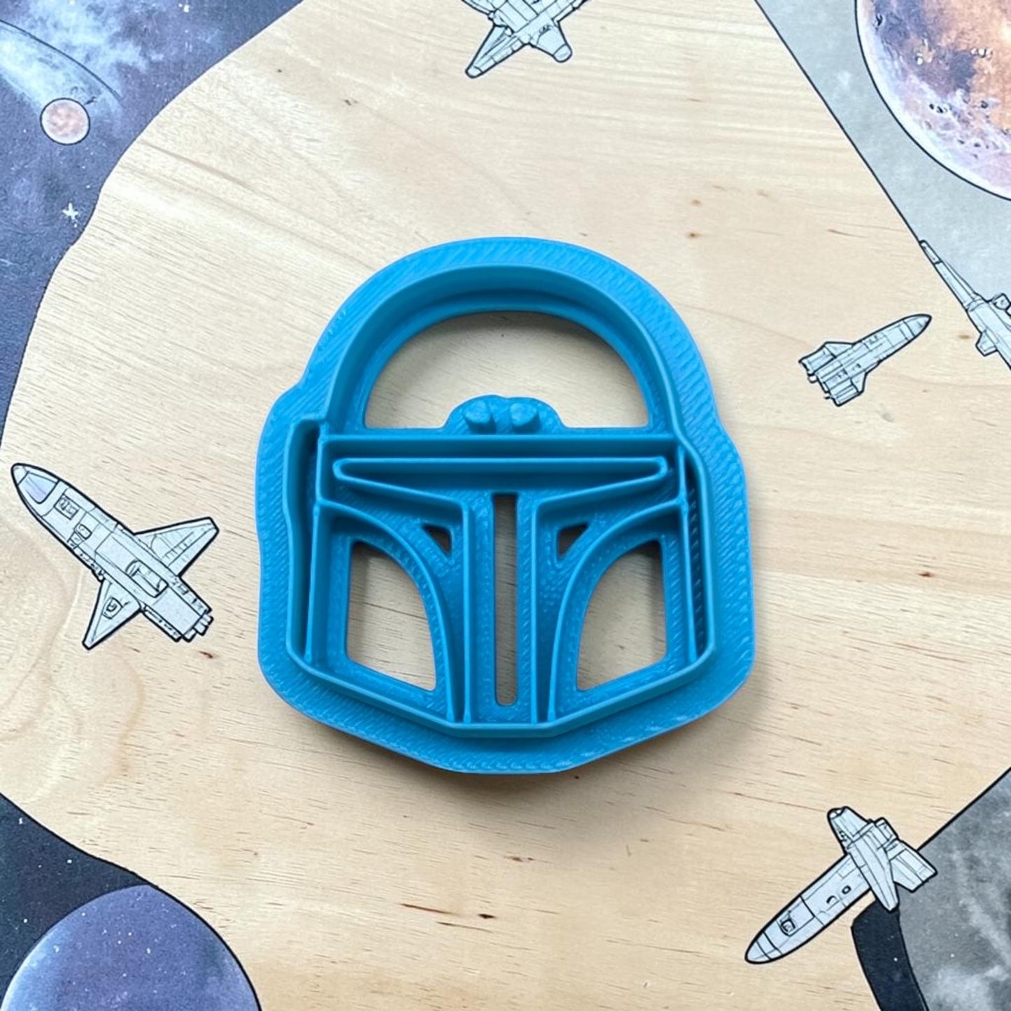 Blue cookie cutter shaped like a helmet on a wooden surface with space-themed illustrations.