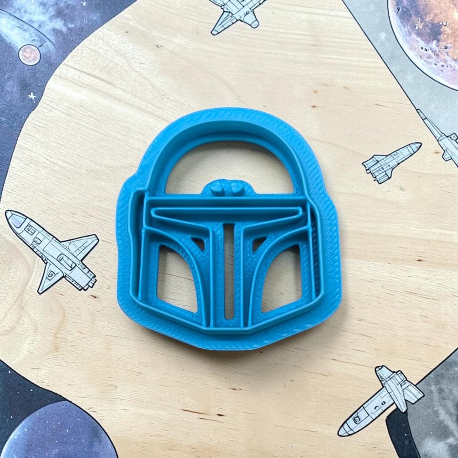 Blue cookie cutter shaped like a helmet on a wooden surface with space-themed illustrations.