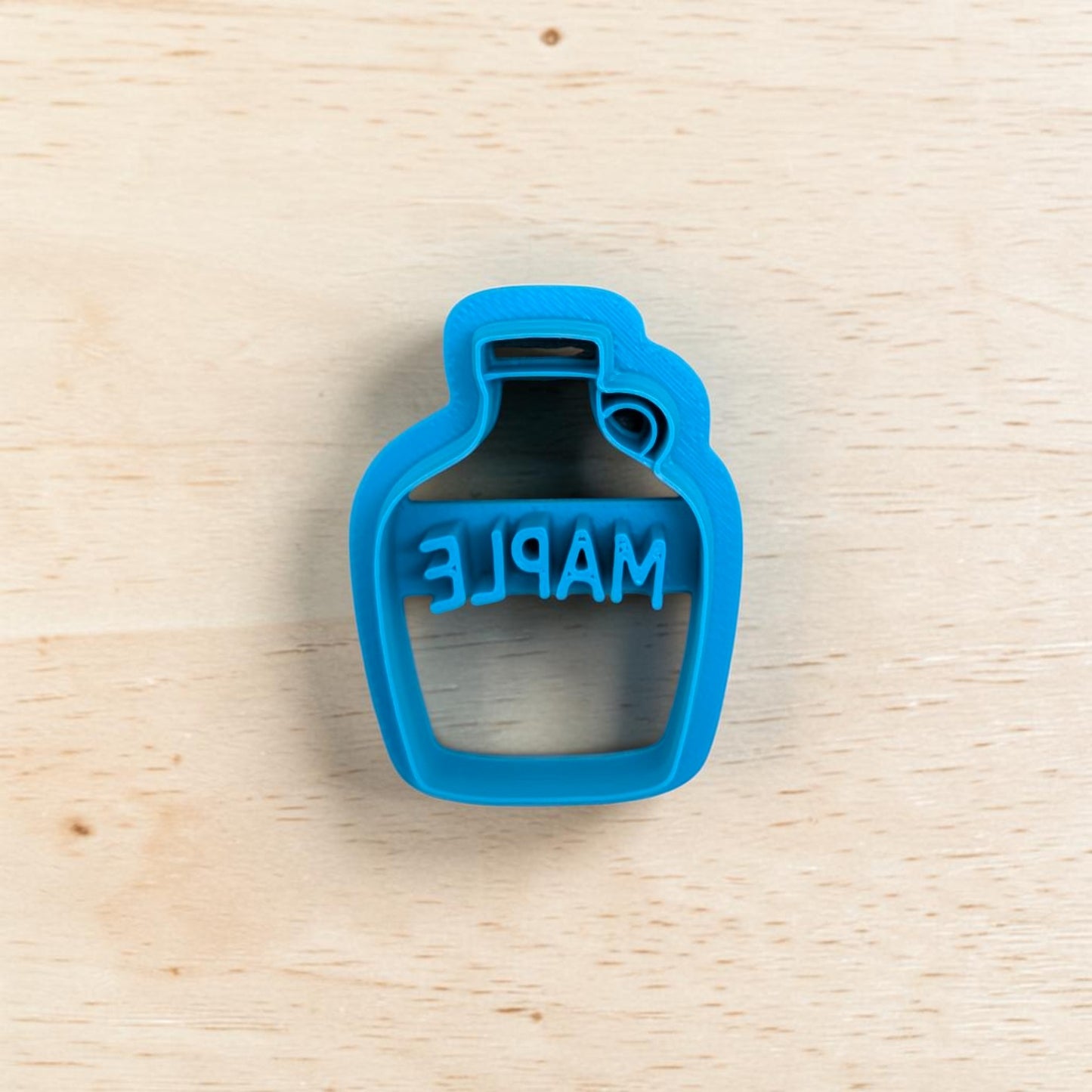 A bright blue plastic bottle-shaped cookie cutter with the word 'MAPLE' embossed, shown on a light wood grain background.