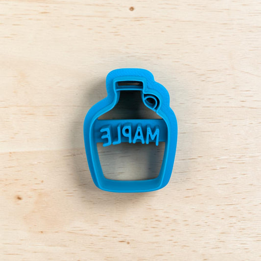 A bright blue plastic bottle-shaped cookie cutter with the word 'MAPLE' embossed, shown on a light wood grain background.