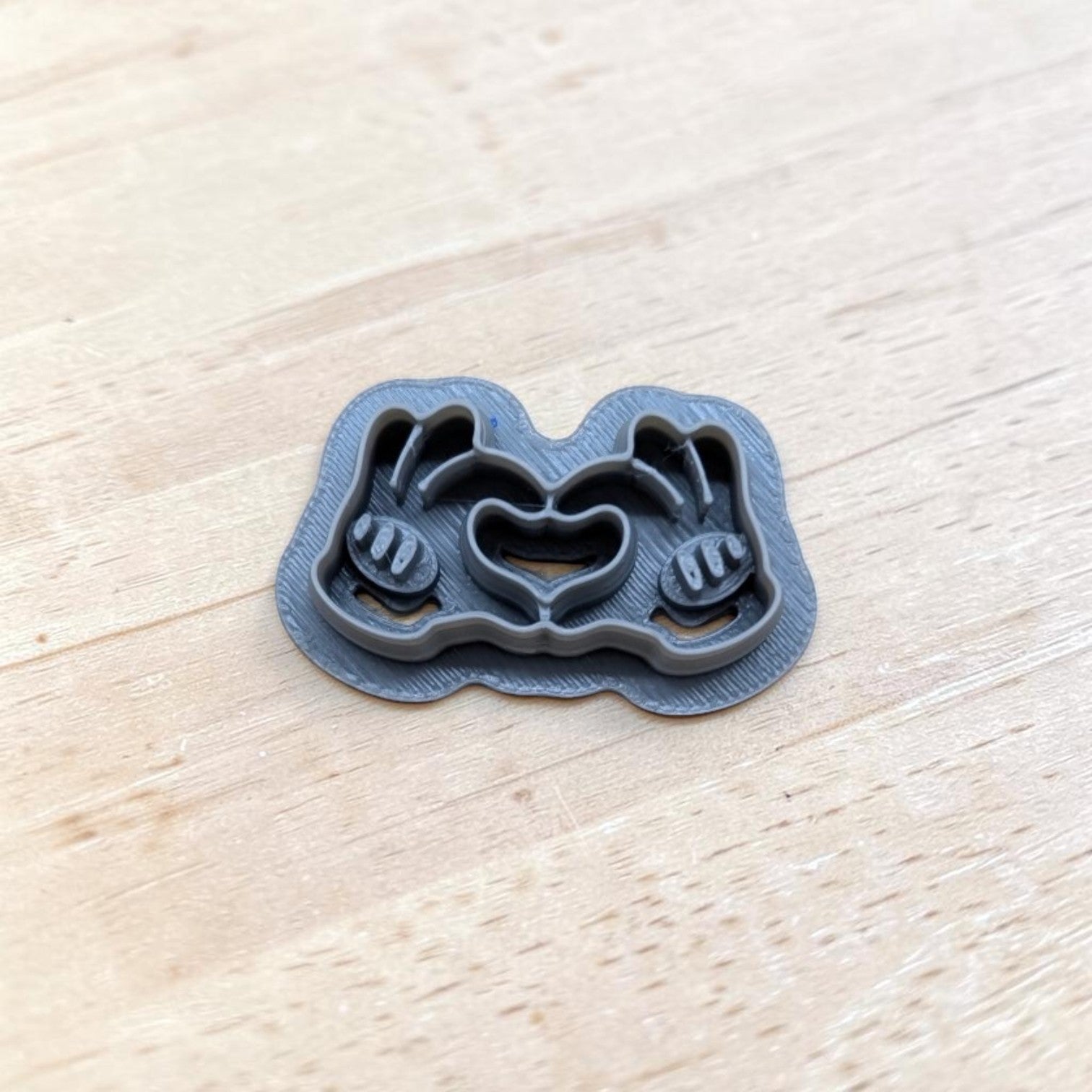 Gray 3D-printed cookie cutter shaped like Mickey Mouse gloved hands forming a heart, displayed on a light wooden surface.