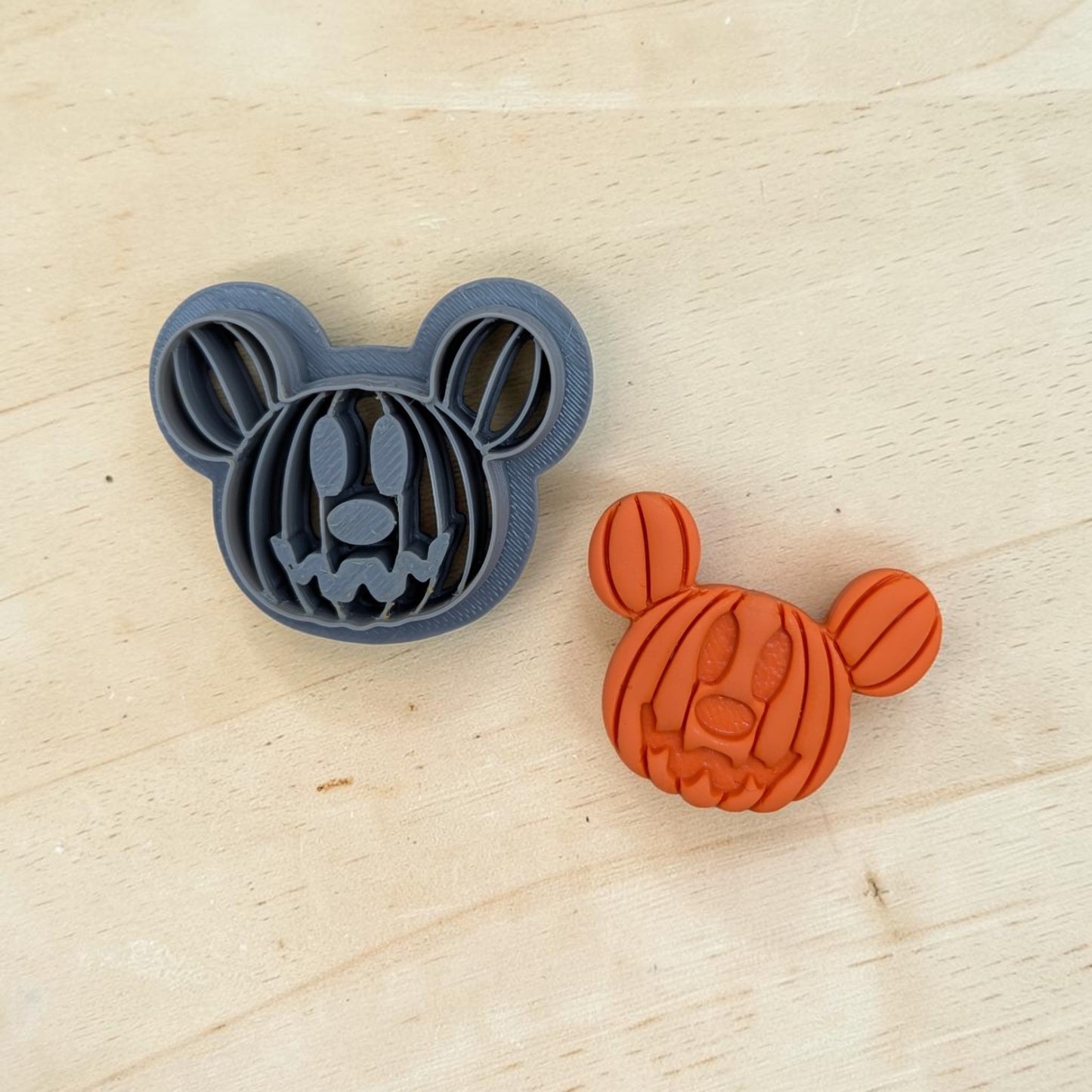 Mickey‑style pumpkin head cookie cutter and orange clay imprint on wood surface, Halloween jack‑o‑lantern design for cookies, fondant, and polymer clay crafts