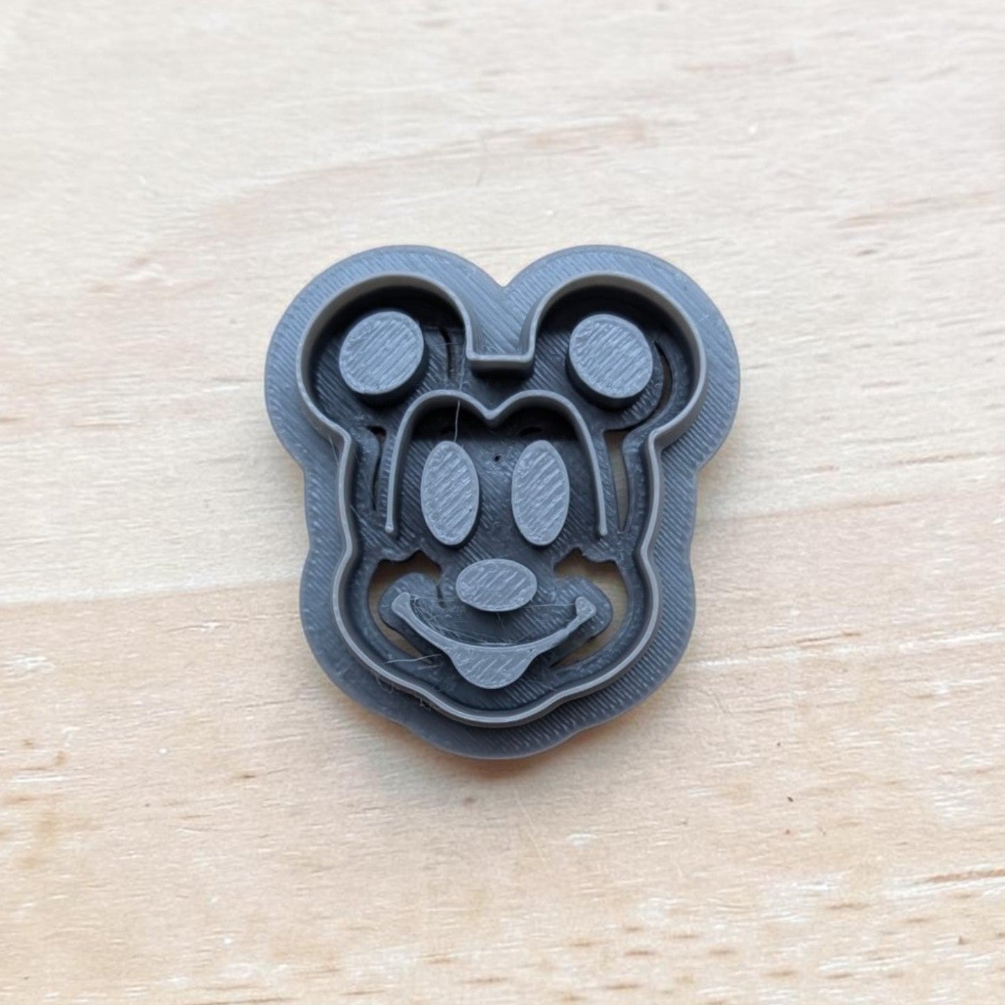 A dark gray 3D-printed Mickey Mouse-shaped cookie cutter or stamp, shown from a top-down view on a light wood grain background.