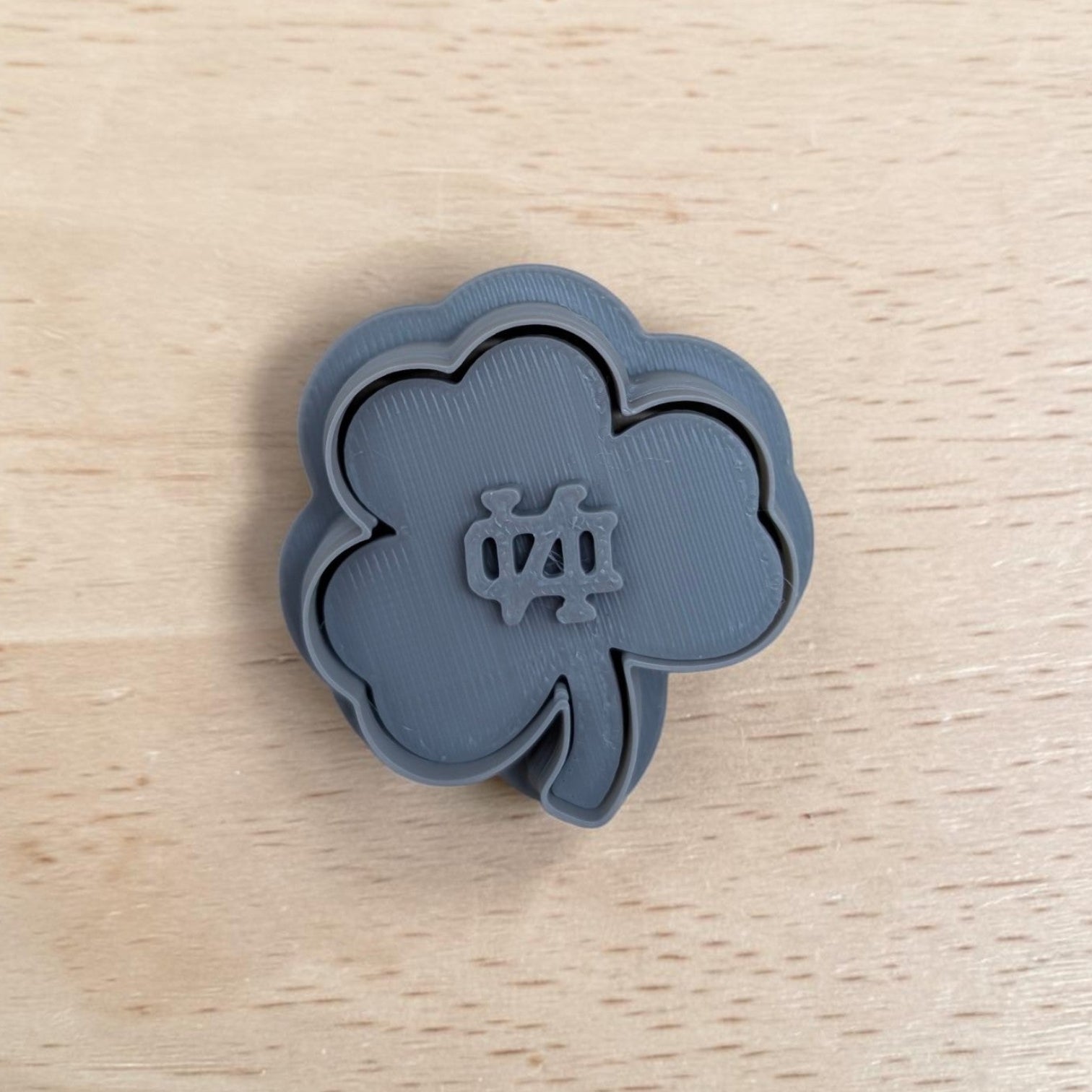 A dark gray 3D-printed shamrock-shaped cookie cutter with a Notre Dame logo in the center, resting on a light wood grain surface.