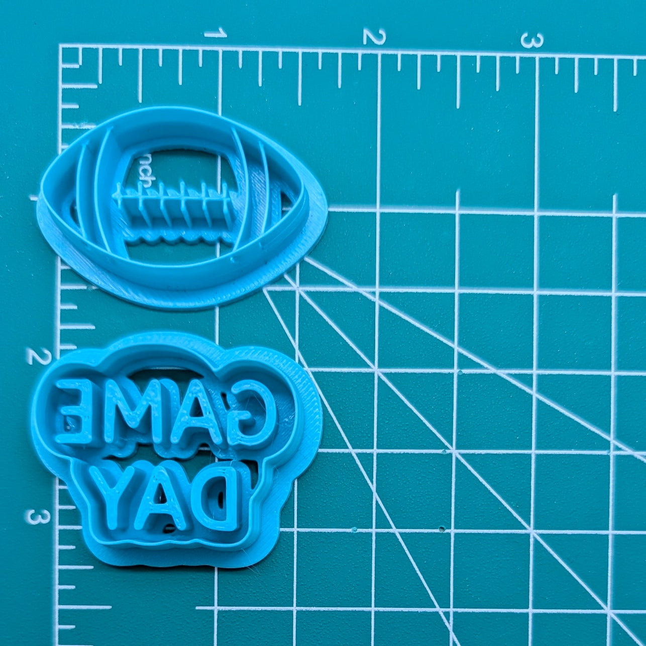A light blue plastic cookie cutter and matching cutter template for a football-shaped cookie, displayed on a teal cutting mat with white grid lines.