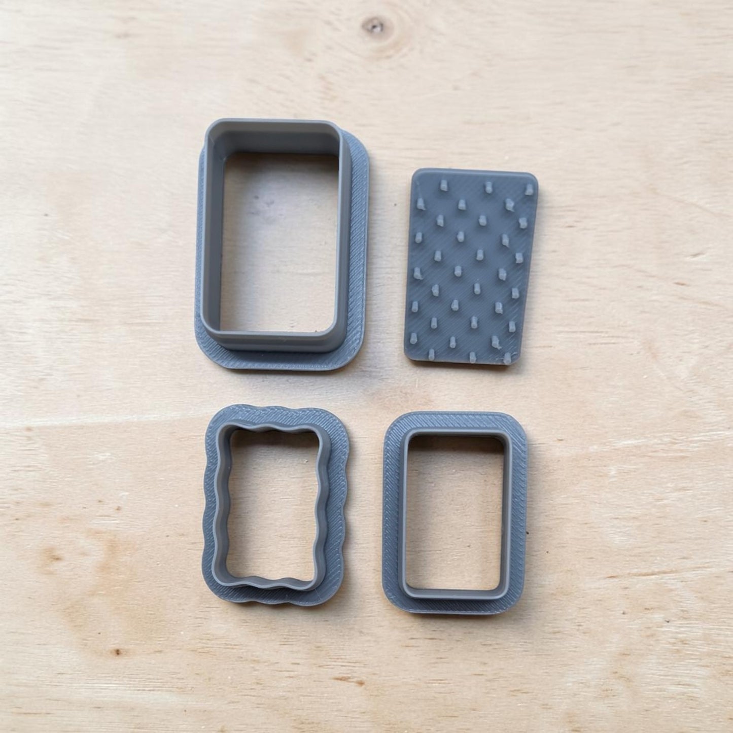 Four gray baking or craft tools, including two rectangular cutters and two textured pieces, arranged on a light wood surface.