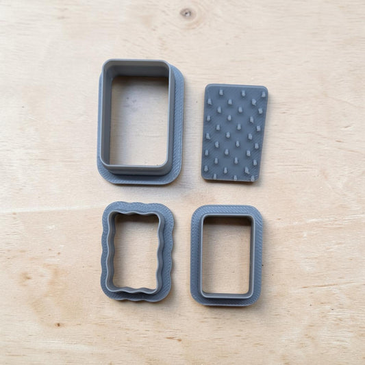 Four gray baking or craft tools, including two rectangular cutters and two textured pieces, arranged on a light wood surface.