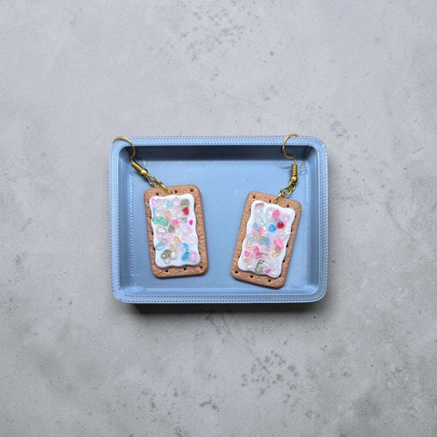 A pair of mini pop tart earrings displayed on a small silver baking tray against a light gray textured background. Each earring has a tan border, a white center filled with small multicolored confetti-like dots, and a gold fishhook ear wire.