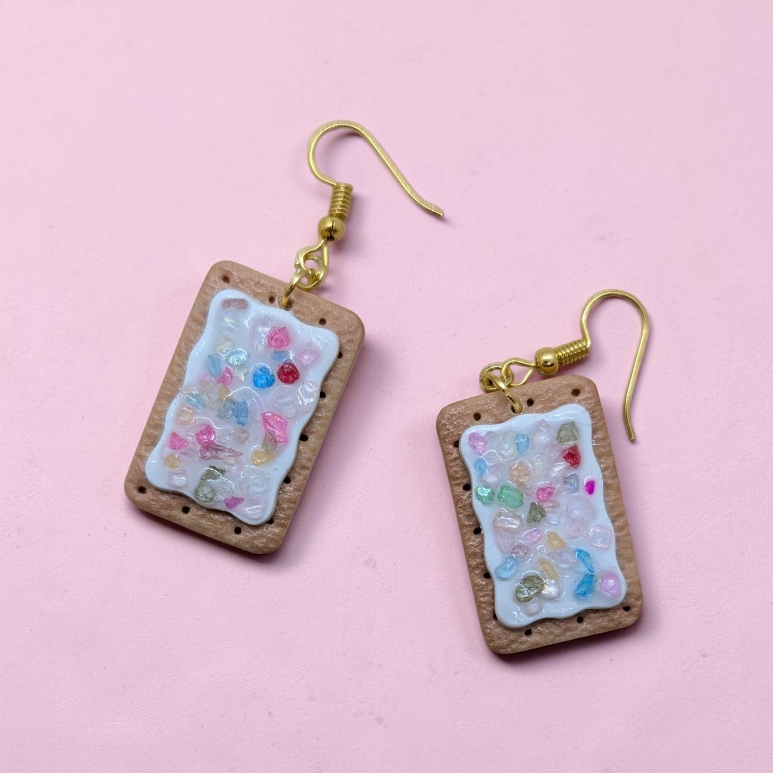 A pair of rectangular dangle earrings shaped like pop tarts with white frosting and colorful candy pieces, hanging from gold fishhook wires on a solid pink background.