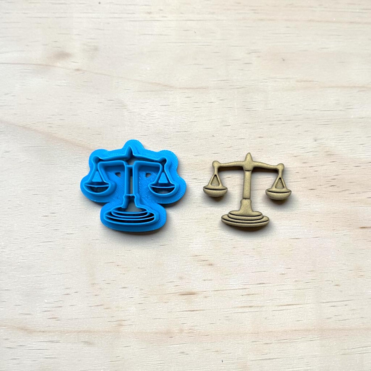 
Scales of Justice cookie cutter and matching metal emblem on light wood background for pottery, clay, fondant, and baking projects.