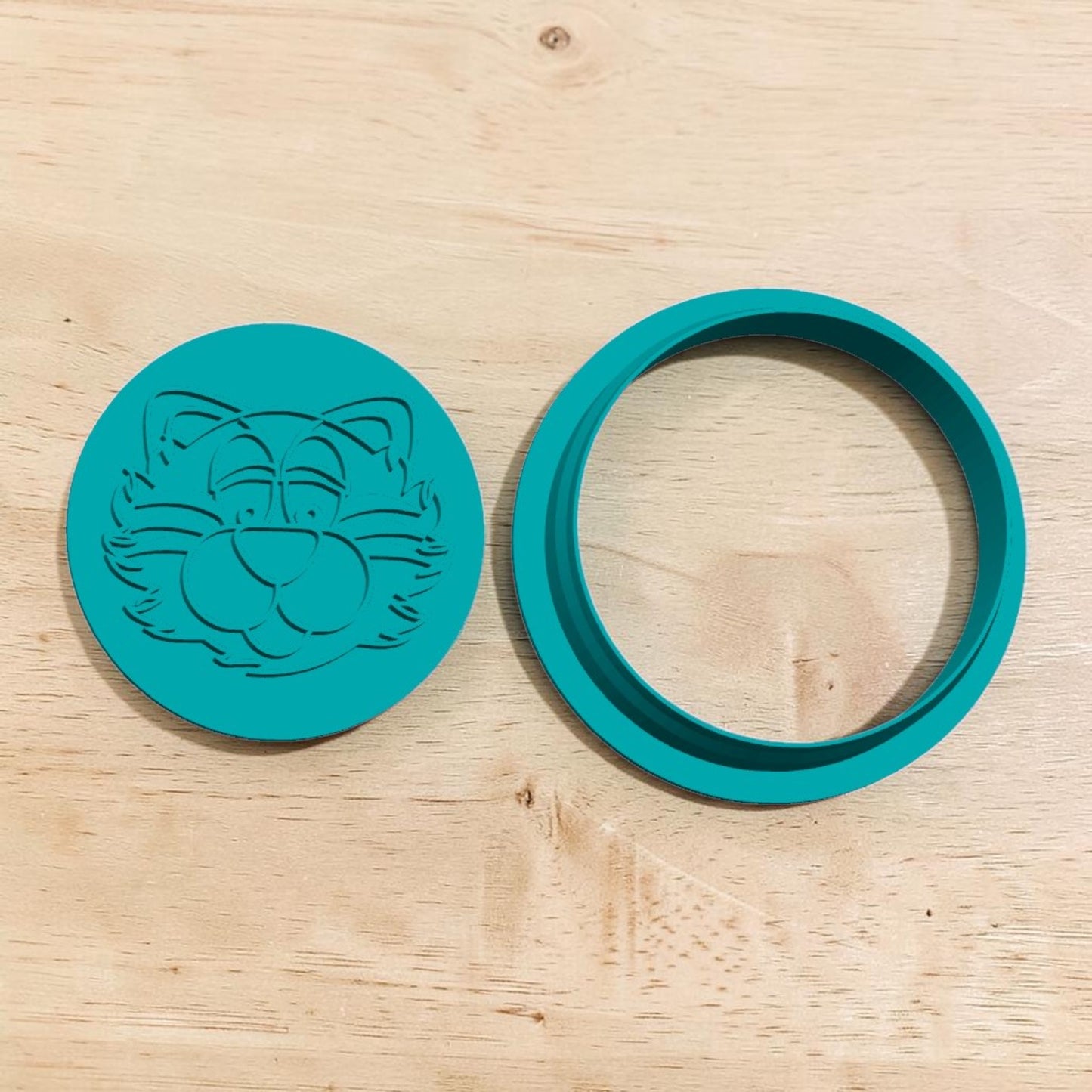 A teal green circular silicone mold or ring set on a light wood grain surface. One piece features an embossed cartoon cat face, and the other is a matching circular ring.
