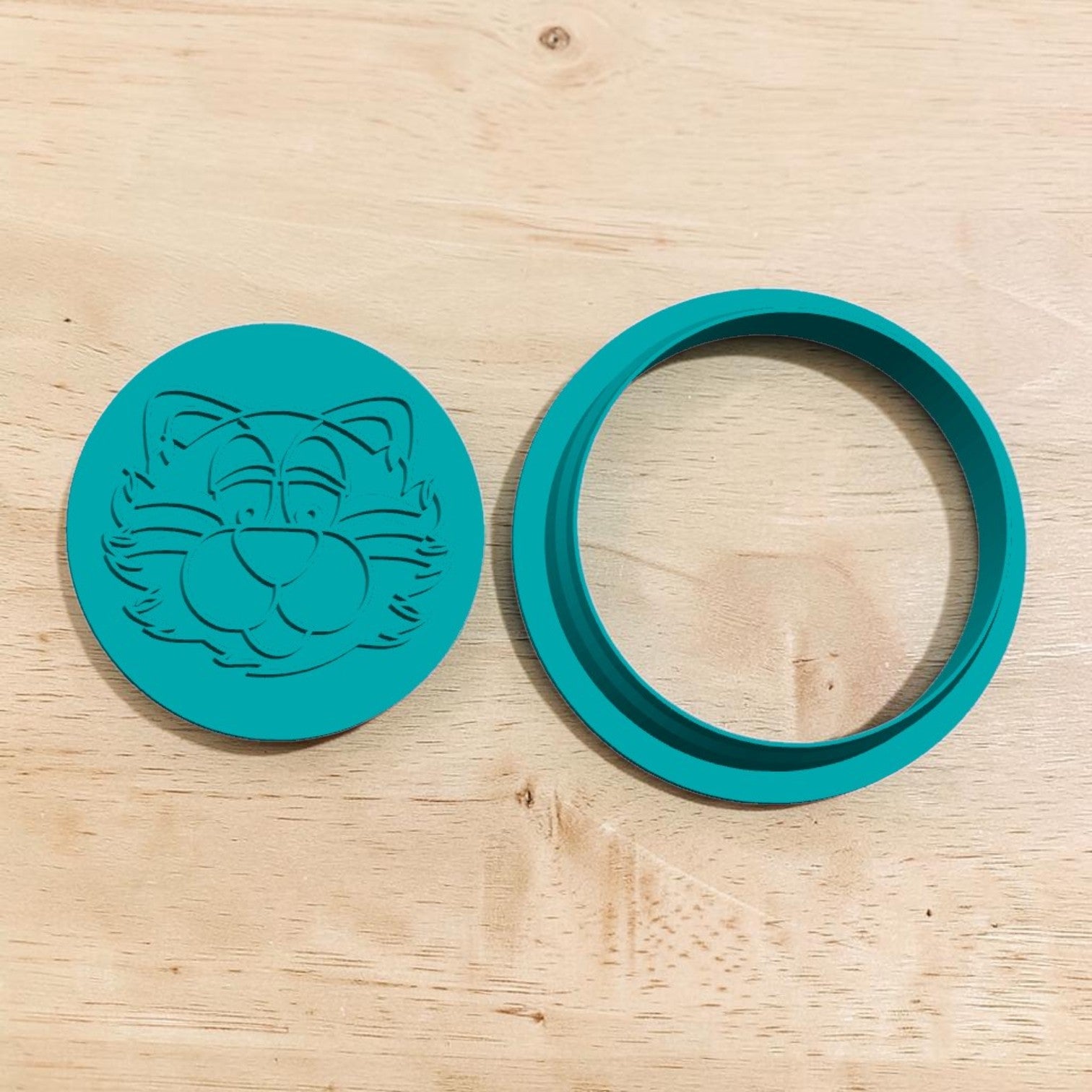A teal green circular silicone mold or ring set on a light wood grain surface. One piece features an embossed cartoon cat face, and the other is a matching circular ring.