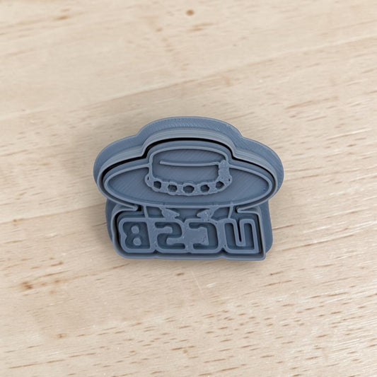 A gray plastic UCSB Gauchos stamp and cutter set, placed on a light wood surface.