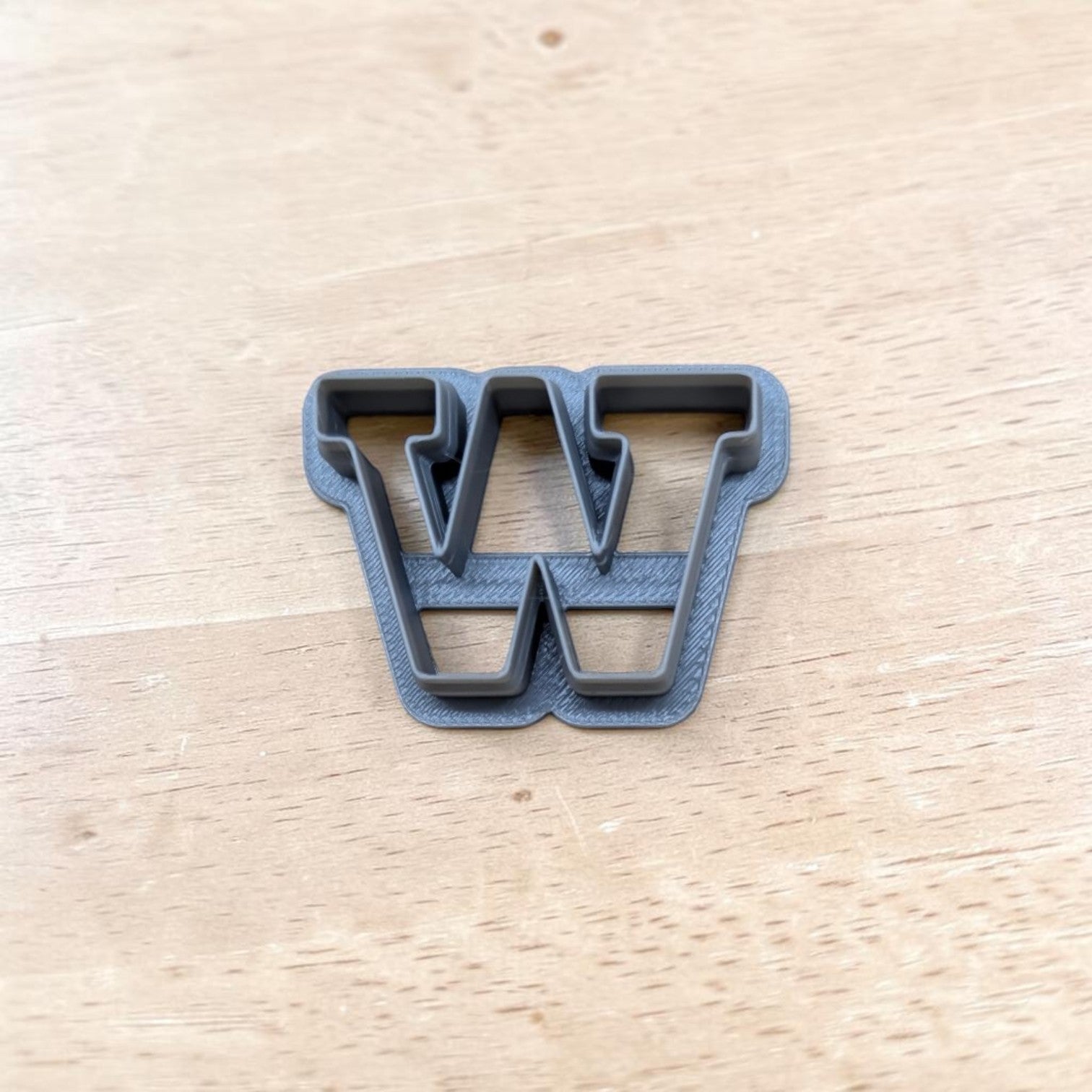 A dark gray plastic letter 'W' cookie cutter resting on a light-colored wooden tabletop.