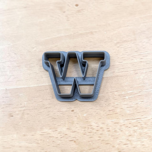 A dark gray plastic letter 'W' cookie cutter resting on a light-colored wooden tabletop.